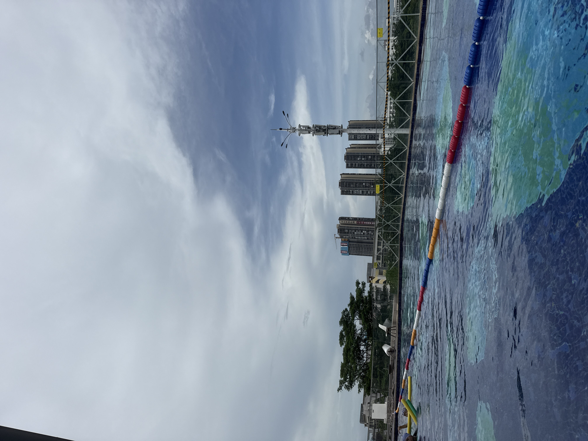 The image depicts a swimming pool with lane markers in the foreground. In the middle ground, there is a fence, some trees, and several buildings, including tall high-rise apartments. A communication tower with multiple antennas is also visible. The background features a cloudy sky with patches of blue. The scene appears to be an outdoor urban setting in Huizhou, China. There are no people actively swimming or interacting within the visible portion of the pool.