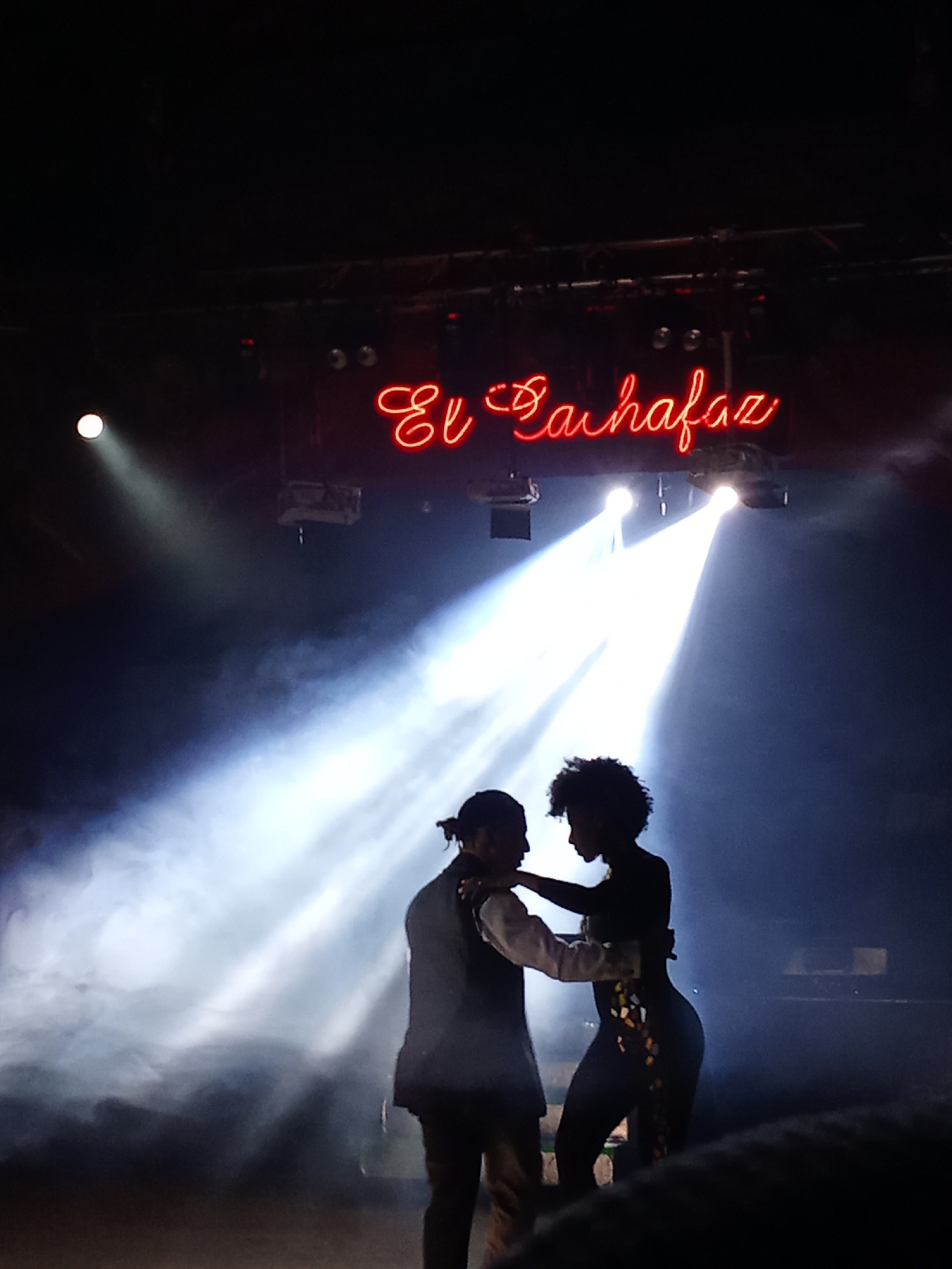 A silhouette of a couple dancing is captured on a dimly lit stage, possibly in the city of Providencia, Chile. The dancers are illuminated by spotlights, creating a dramatic effect with visible smoke or haze in the air. Above them, a neon sign in red cursive reads "El Cachafaz". The male dancer is on the left, facing the female dancer who is on the right. The female dancer is wearing a dress with what appears to be colorful embellishments. The stage lighting is intense, with beams cutting through the smoky atmosphere.