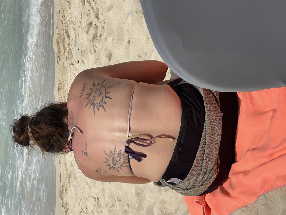 An individual is depicted from behind, seated on a sandy beach at Kendwa Beach, Zanzibar, Tanzania, facing the ocean. The specific location is indicated by coordinates 6° 43' 01" S 39° 11' 18" E tattooed on the individual's right upper back. The individual has dark brown hair secured in a bun and exhibits reddened skin on the upper back, consistent with sunburn. Attire includes a multi-colored bikini top (purple, white, pink, beige straps visible) tied in the back, black swim bottoms, and a grey and brown textured garment wrapped around the waist. An orange beach towel is positioned beneath the individual. Tattoos on the back include an Eye of Ra/Horus symbol within a sun on the left shoulder blade, a sun and crescent moon symbol with a face on the right shoulder blade, and text "La Froufrousse" with "PARADISE 2021" visible below it on the upper back. The immediate environment consists of pale sand, with turquoise ocean water and waves in the background under bright daylight. A partial grey, curvilinear object occupies the lower right foreground.