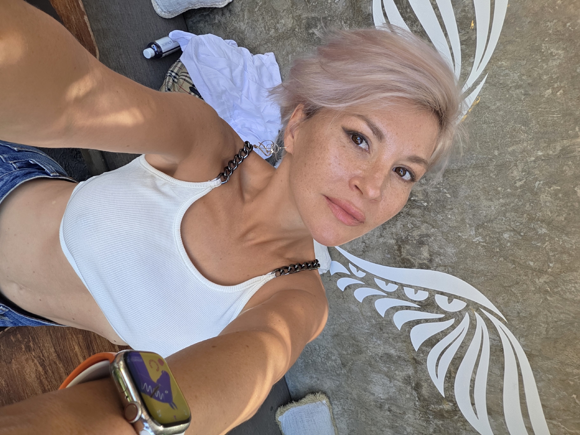 A light-skinned woman with short, dyed blonde hair is taking a selfie from a high angle. She is wearing a white tank top with a chain strap, and a smartwatch with an orange strap is visible on her left wrist. She is standing in front of a textured gray surface with a white design resembling a wing. A white shirt and a small bottle are visible in the upper left corner of the frame. The photograph was taken in North Kuta, Indonesia.