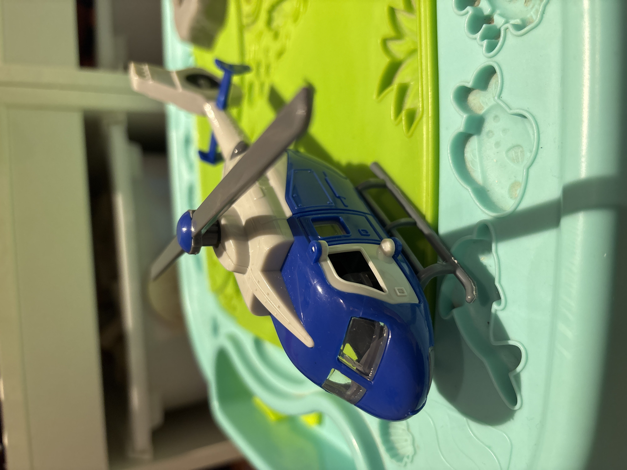 A blue and white toy helicopter is positioned on a bright green surface, which is itself resting on a mint green play tray. The helicopter has two rotors, a cockpit with windows, and landing gear. The play tray has molded shapes resembling water or clouds. The background is blurred but appears to be shelving units. The lighting suggests an indoor setting, likely a home. There are no people or animals visible in the image. The scene does not provide any information that could indicate its location in Dubai, United Arab Emirates.
