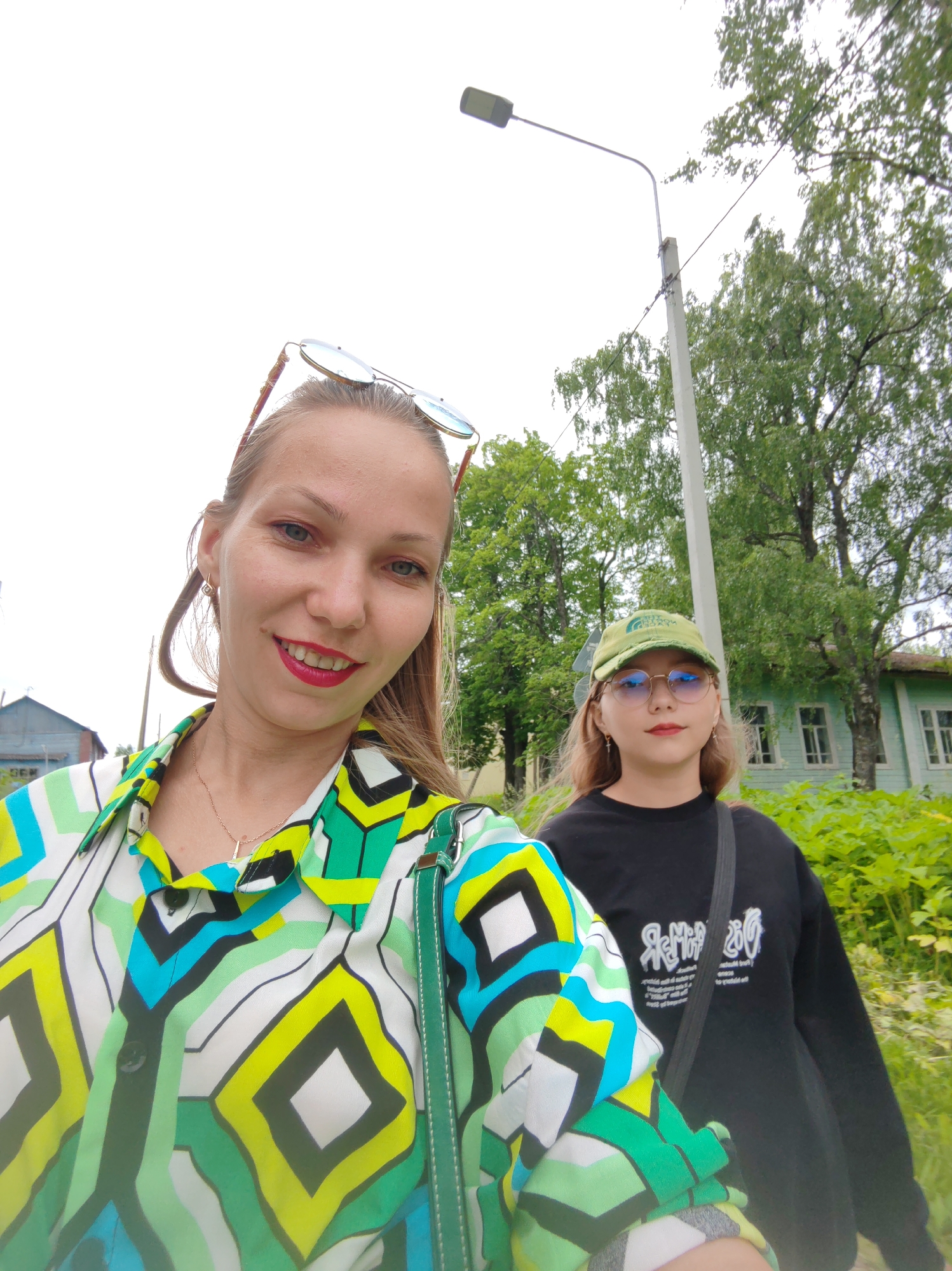 Two women, one in the foreground and another behind her, are taking a selfie outdoors. The woman in the foreground is wearing a colorful, geometric-patterned shirt and has sunglasses pushed up on her head. The woman behind her is wearing a black sweatshirt and a green baseball cap, also with sunglasses. They are standing in front of a light-colored building with green trim, surrounded by lush green foliage and trees. A street lamp and a utility pole are visible in the background. The sky is overcast. The location is identified as Chukhloma, Russia.