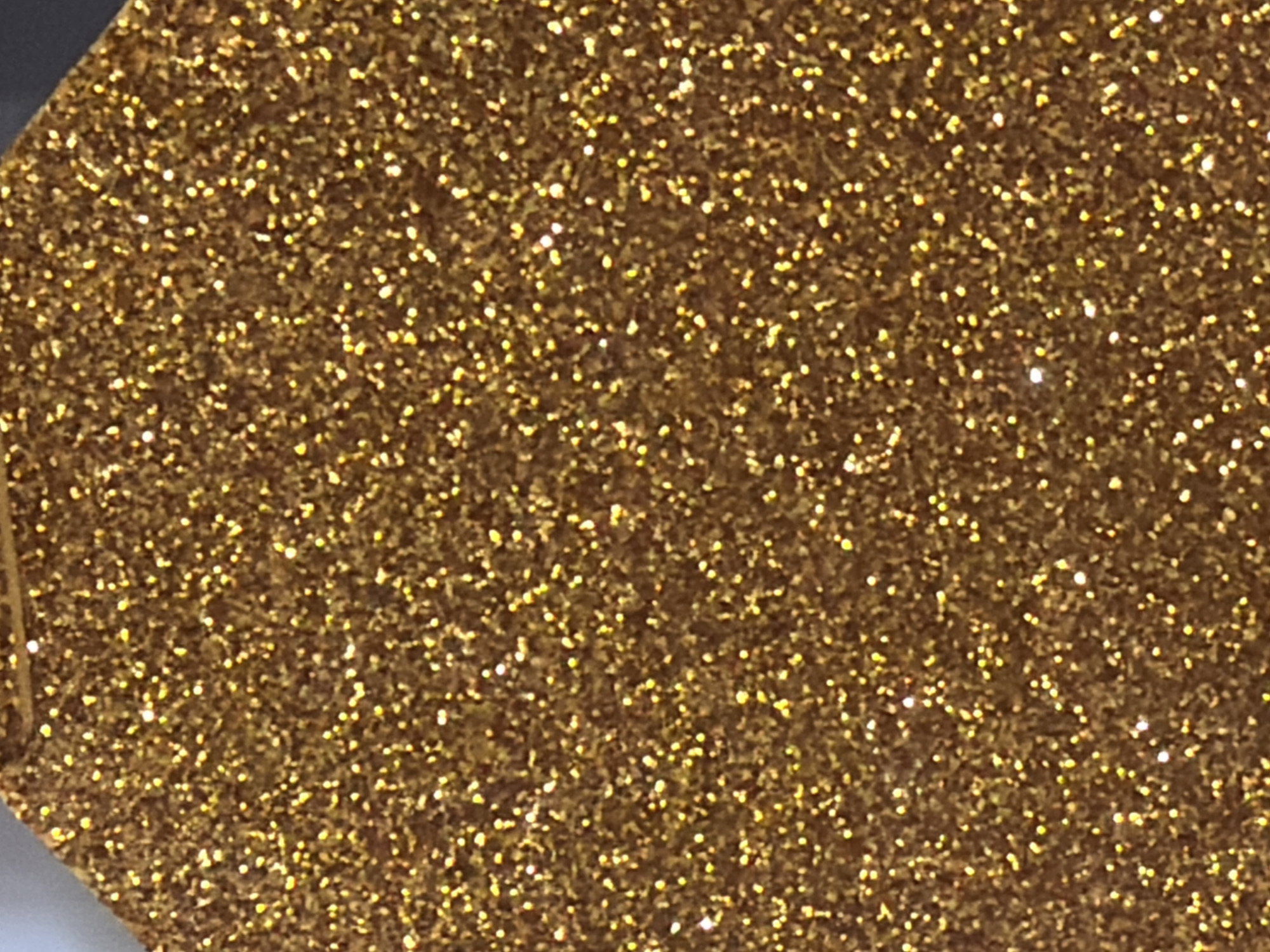 The image is a close-up, overhead shot of a textured surface covered in gold glitter. The glitter appears to be applied to a dark base material, forming a dense, sparkling pattern. The lighting highlights the reflective qualities of the glitter, creating numerous small points of light across the surface. The image is abstract and does not contain any discernible elements of a cityscape, environment, people, or actions. Therefore, it is not possible to provide details about the location in the city of Arcahueja, Spain, as the image does not depict such a setting.