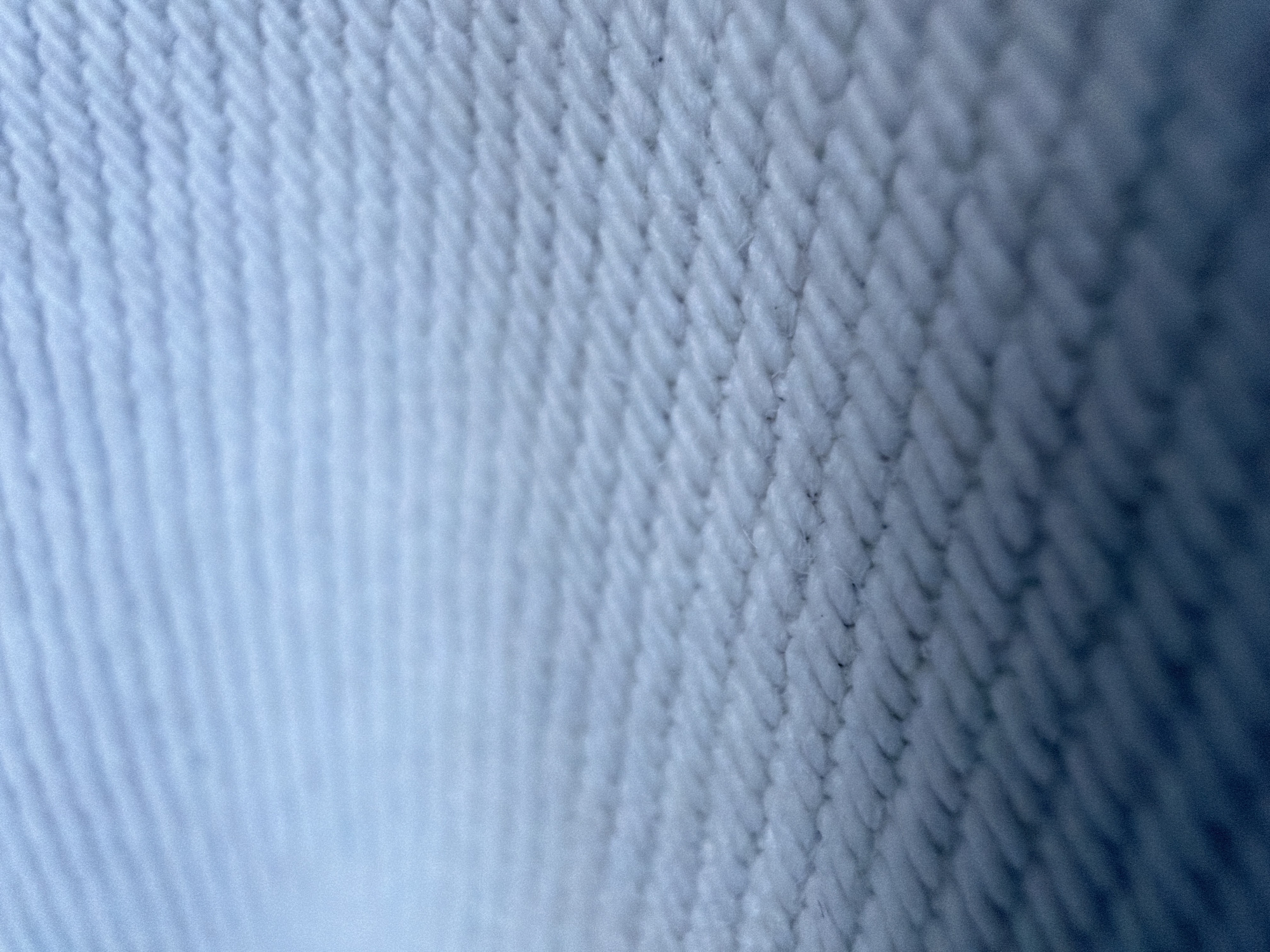 This media file is a close-up, abstract photograph of a textured blue fabric. The fabric appears to be woven, with a pattern of interlocking threads creating a ribbed or corded surface. The lighting creates shadows and highlights that emphasize the texture, giving the fabric a three-dimensional quality. The left side of the image is softly blurred and fades into a lighter blue, suggesting depth and a light source from that direction. The right side of the image is sharper and darker, revealing the intricate weave of the fabric more clearly. There is no discernible subject, activity, or setting beyond the fabric itself. No text is visible. The overall impression is one of detail and texture, with a cool color palette.