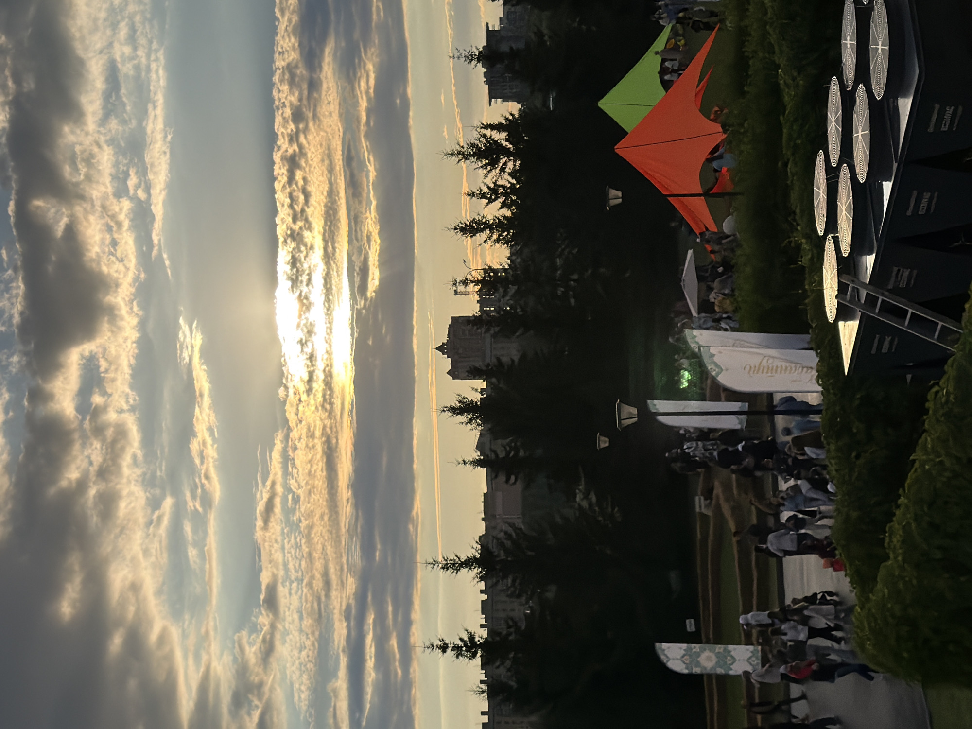 The image shows a city park scene at sunset. The sky is filled with dramatic clouds and rays of sunlight break through. In the foreground, there are large, dark HVAC units with circular fans on top. A ladder leans against one of the units. Further back, there is a group of people walking on a paved path, some standing around and talking. Colorful tents, one orange and one green, are set up in the park. Evergreen trees are silhouetted against the sky, and buildings are visible in the distance. Several tall, thin flags with decorative patterns are also present in the park. The overall atmosphere suggests an outdoor event or gathering.