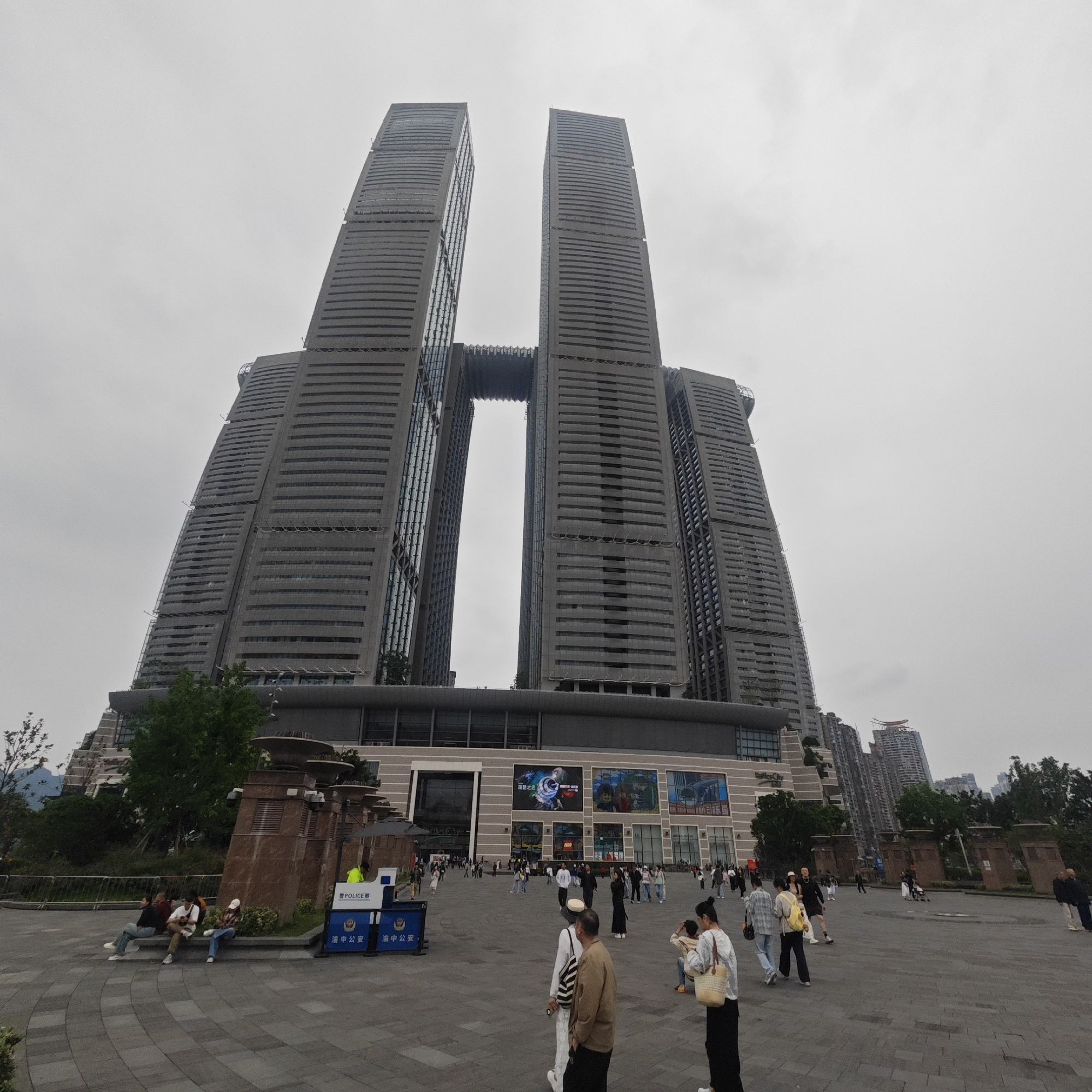 The image shows a wide-angle view of Raffles City Chongqing in Yuzhong District, China, on an overcast day. The complex is composed of eight towers connected by a sky bridge, a large shopping mall, and various other commercial spaces.
In the foreground, a paved plaza is populated with pedestrians, some walking, others standing or sitting. Several people are carrying bags or holding children. A police barricade is visible to the left, indicating a security presence.
The backdrop is the complex itself, dominated by the towers and sky bridge. The facade of the shopping mall below features large advertising displays. The surrounding buildings of Yuzhong District are visible in the distance.