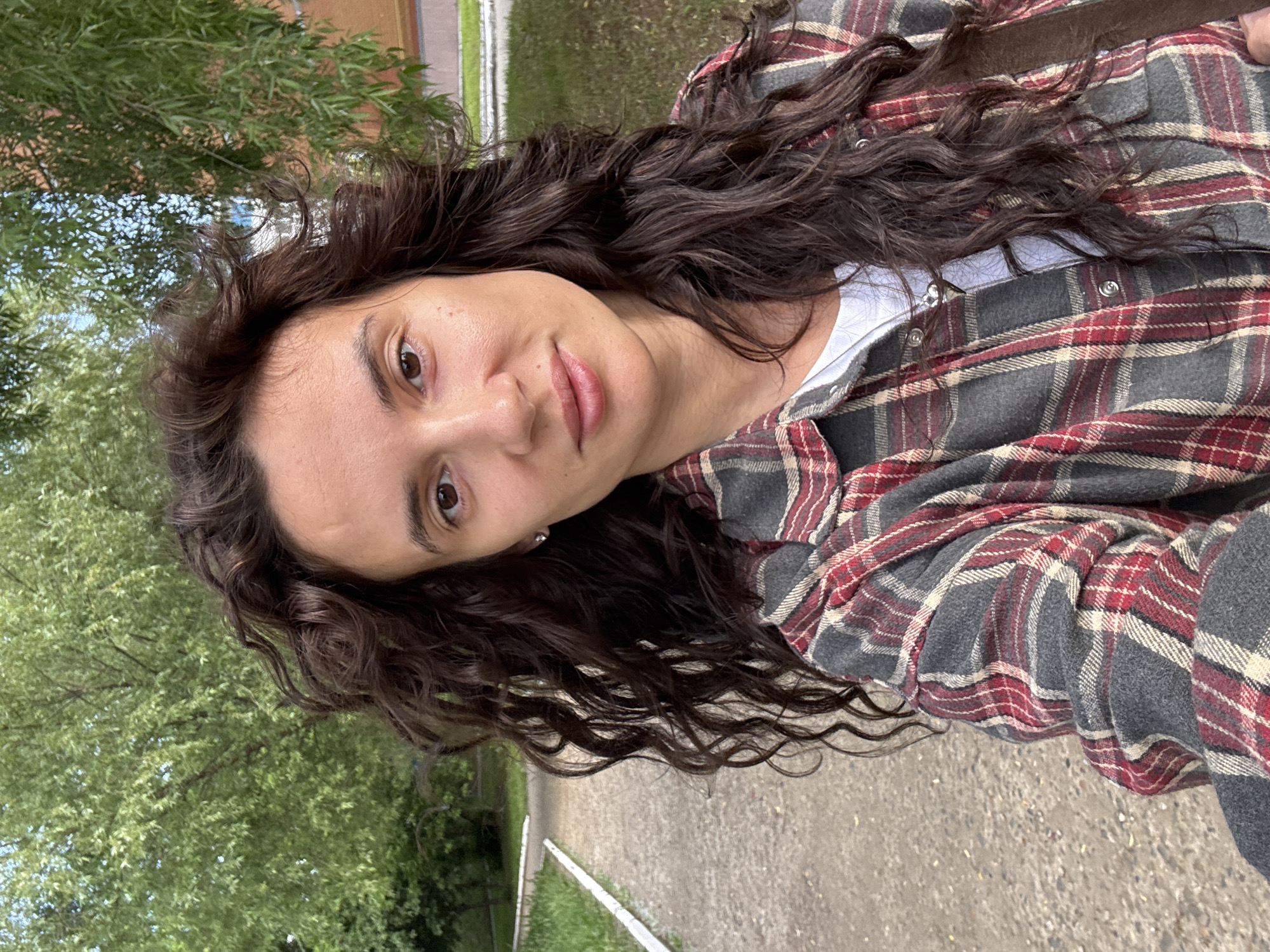 A selfie-style portrait captures a young woman outdoors in what appears to be Chaikovsky, Russia. The woman, with long, dark, wavy or curly hair, is wearing a red, grey, and off-white plaid flannel shirt unbuttoned over a white t-shirt. Her face is turned towards the camera, with her head tilted at a significant angle, looking directly into the lens. She has brown eyes and full lips, with a neutral to slightly contemplative expression.
The background shows lush green trees and foliage, suggesting a natural outdoor setting like a park, garden, or a tree-lined street. A paved path or road is visible behind her, with glimpses of what might be a light-colored fence or curb line. The natural lighting indicates it is daytime, and the vibrant green of the leaves suggests clear weather. No specific activities are depicted beyond the act of taking the photo itself. There is no visible text in the image.