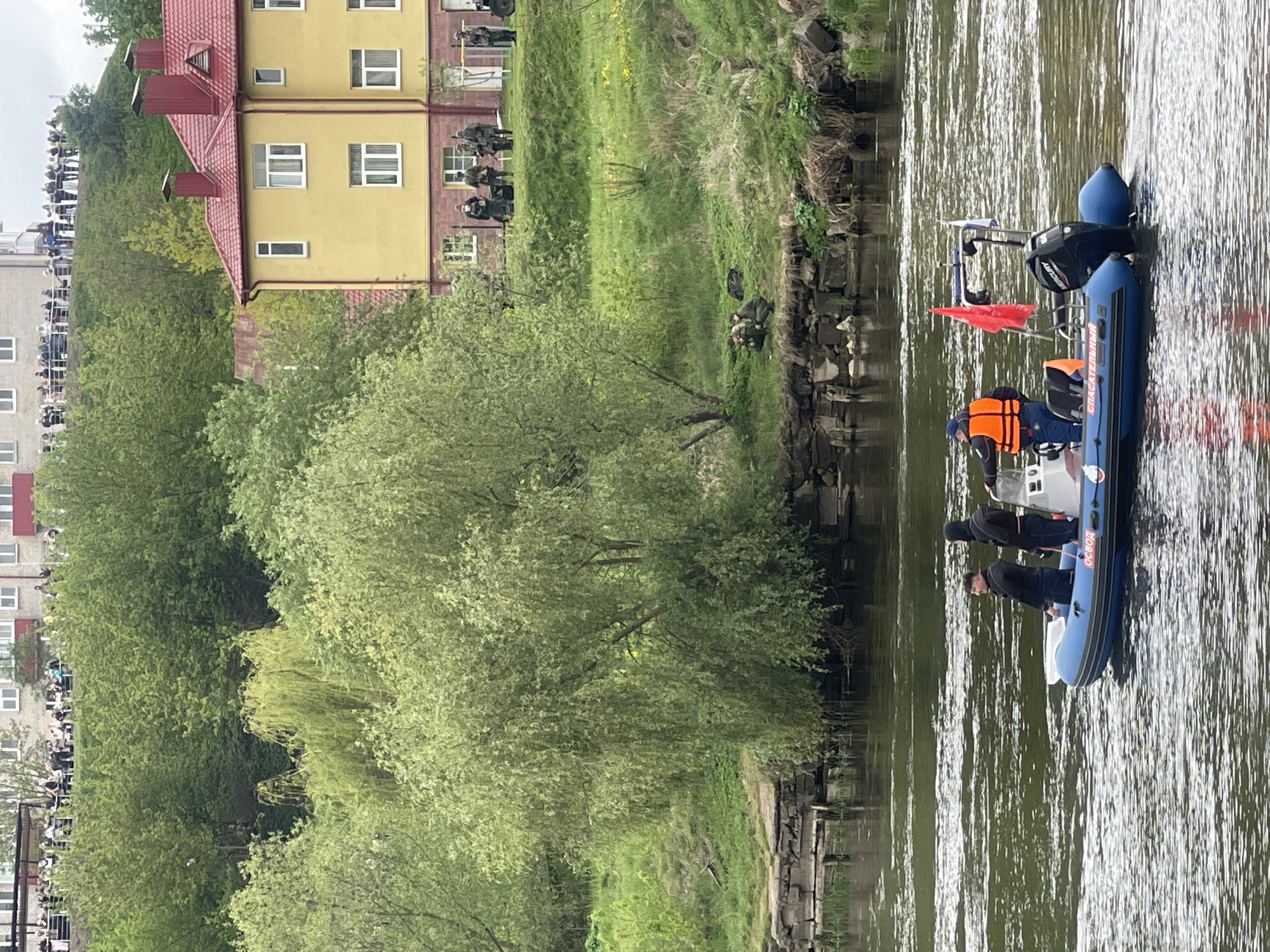 The image captures a scene along the Neman River in Hrodna, Belarus. A blue inflatable rescue boat, labeled "СПАСАТЕЛЬНЫЙ" (Rescue), with two occupants wearing life vests, floats on the water. A Mercury outboard motor is attached to the boat. On the riverbank, people in dark tactical clothing appear to be conducting a search operation. Above them, a yellow building with a red roof and windows is visible. Further away, a crowd of onlookers gathers along a higher vantage point. Lush green vegetation lines the riverbank.
