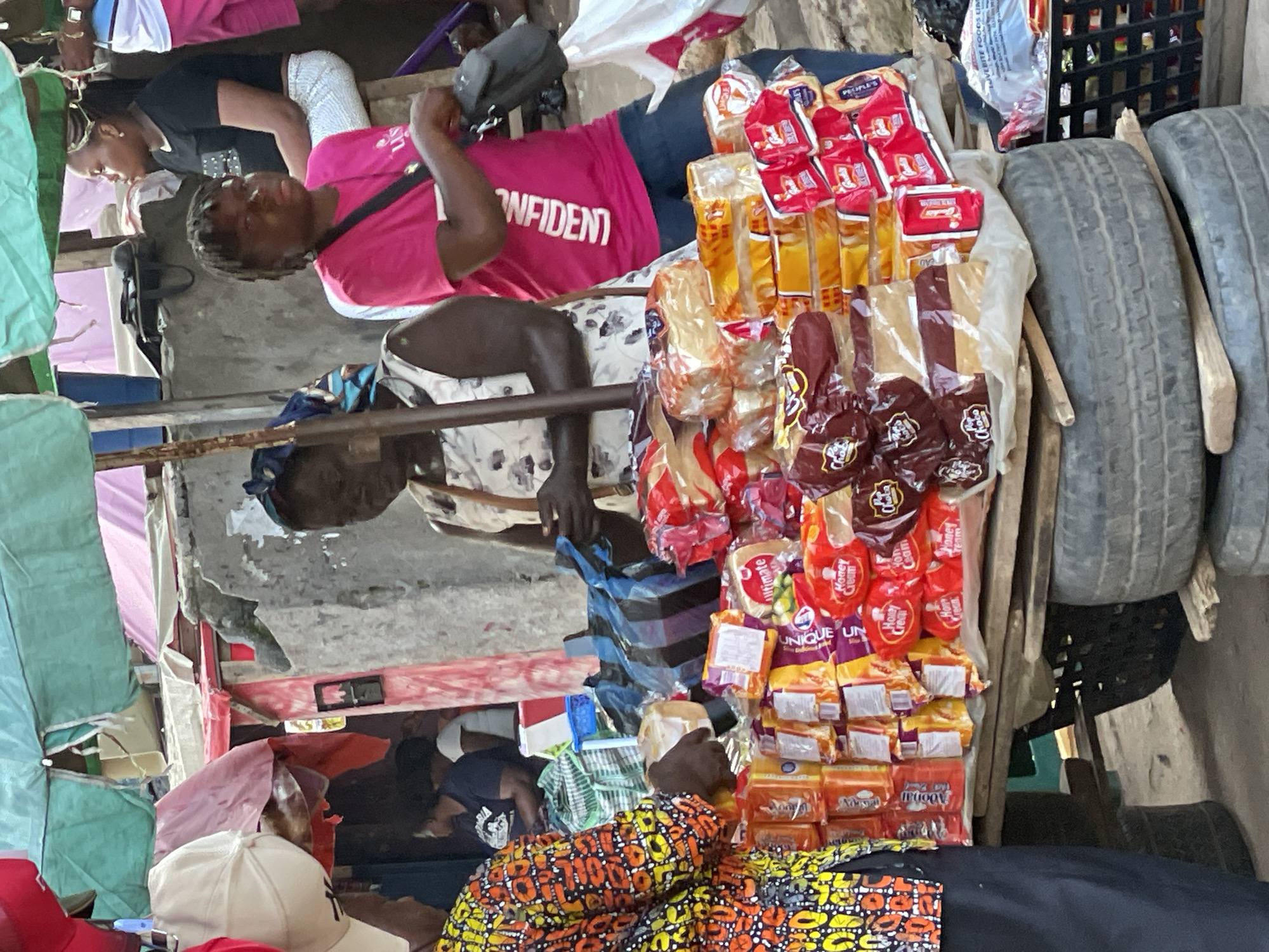 A street market scene in Nkpor, Nigeria, features a display of packaged bread loaves on a wooden cart made from stacked tires. The bread is arranged in rows and columns, with some loaves visible in clear plastic packaging showing brown bread and others in red and yellow wrappers. Behind the bread, a woman wearing a bright pink t-shirt with the word "CONFIDENT" printed vertically in white and dark blue jeans stands with a black crossbody bag. To her left, another woman in a white dress with a floral pattern is partially visible, appearing to interact with the bread display. In the background, a concrete wall and various stalls with awnings in green and pink are present. Other people are faintly visible in the background. The overall environment suggests a busy outdoor market setting.