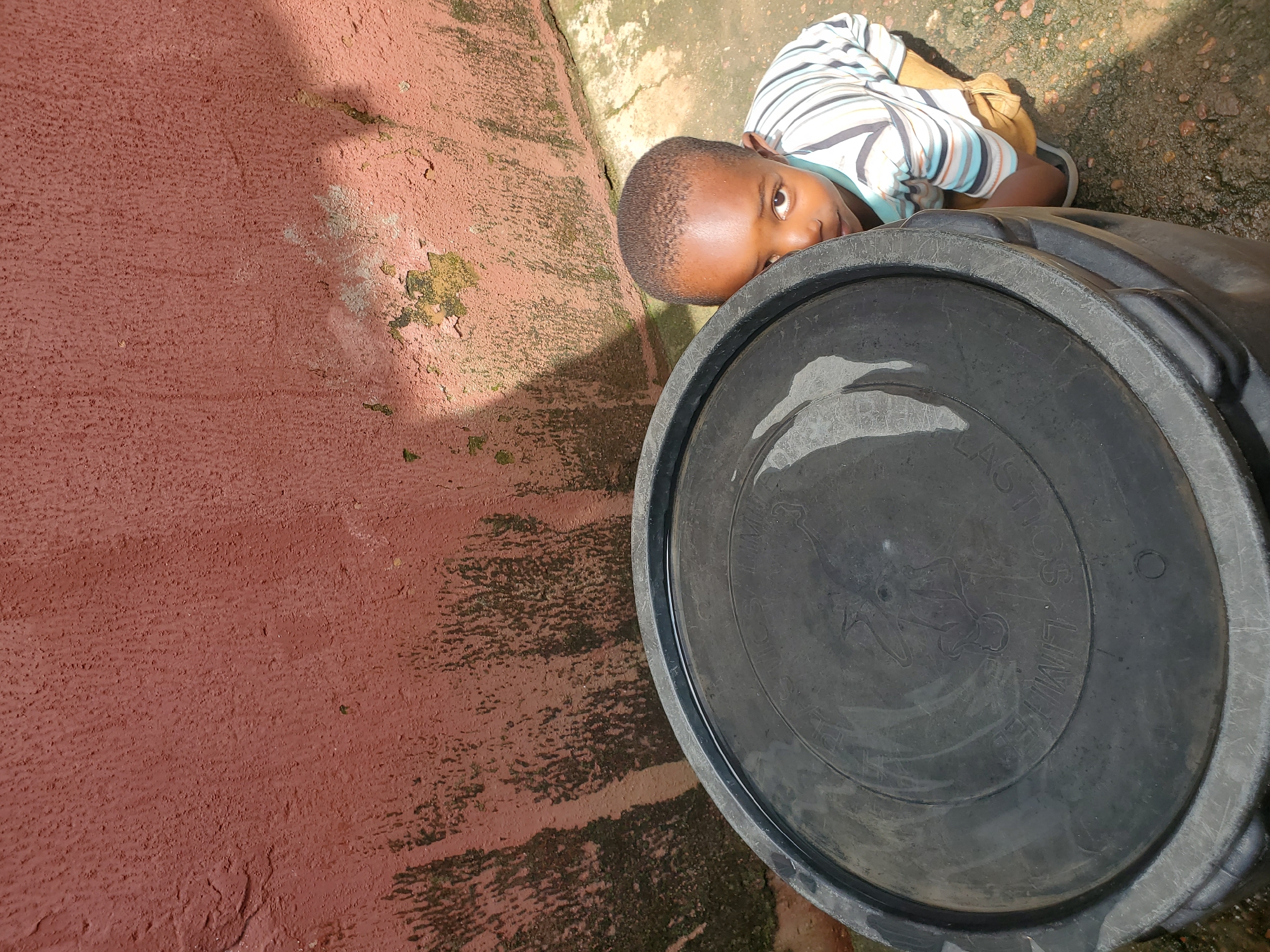 A young child is peeking over a large, black plastic water tank. The child is wearing a striped t-shirt with blue, white, and brown stripes. They have short, dark hair and are looking directly at the camera with a curious expression. The child is lying down, with their head and upper body visible. The water tank appears to be outdoors, resting on a concrete or paved surface.
The background consists of a reddish-pink textured wall on the left and the ground on the right. The ground is a mix of concrete and dirt, with some patches of moss or algae, suggesting an outdoor environment that may be damp. Sunlight is casting shadows on the wall and the ground, indicating it is daytime, likely with bright sunlight. There is some wear and tear visible on the wall, with patches of peeling paint and discolored areas.
The lid of the water tank has some embossed text and a circular design, though it is difficult to read clearly. One visible word appears to be "LIMITED." The scene evokes a sense of everyday life in a warm climate, possibly in a residential or communal area. The image is shot from a high angle, looking down at the child and the tank. The overall mood is candid and captures a moment of observation.