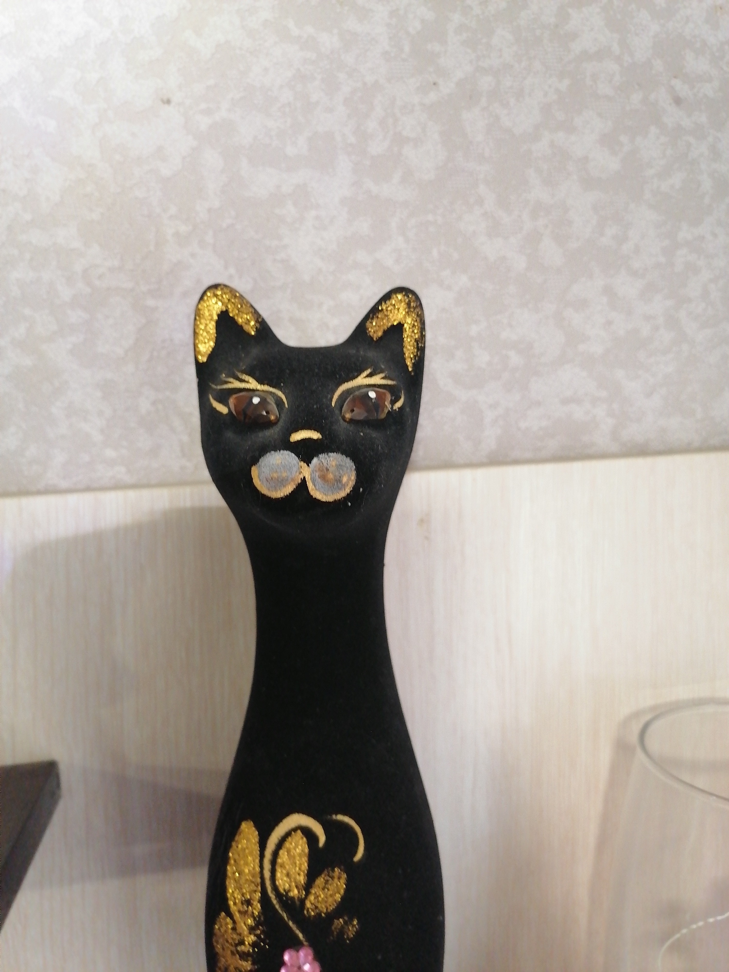 A black velvet cat figurine is shown against a light-colored, textured wall. The figurine has gold glitter on its ears and gold painted accents around its eyes, nose, and mouth. It also has decorative gold swirls and a pink flower on its chest. The figurine is positioned centrally in the frame, with a blurry glass visible to the right. The overall scene suggests an indoor setting, likely a home or shop display.