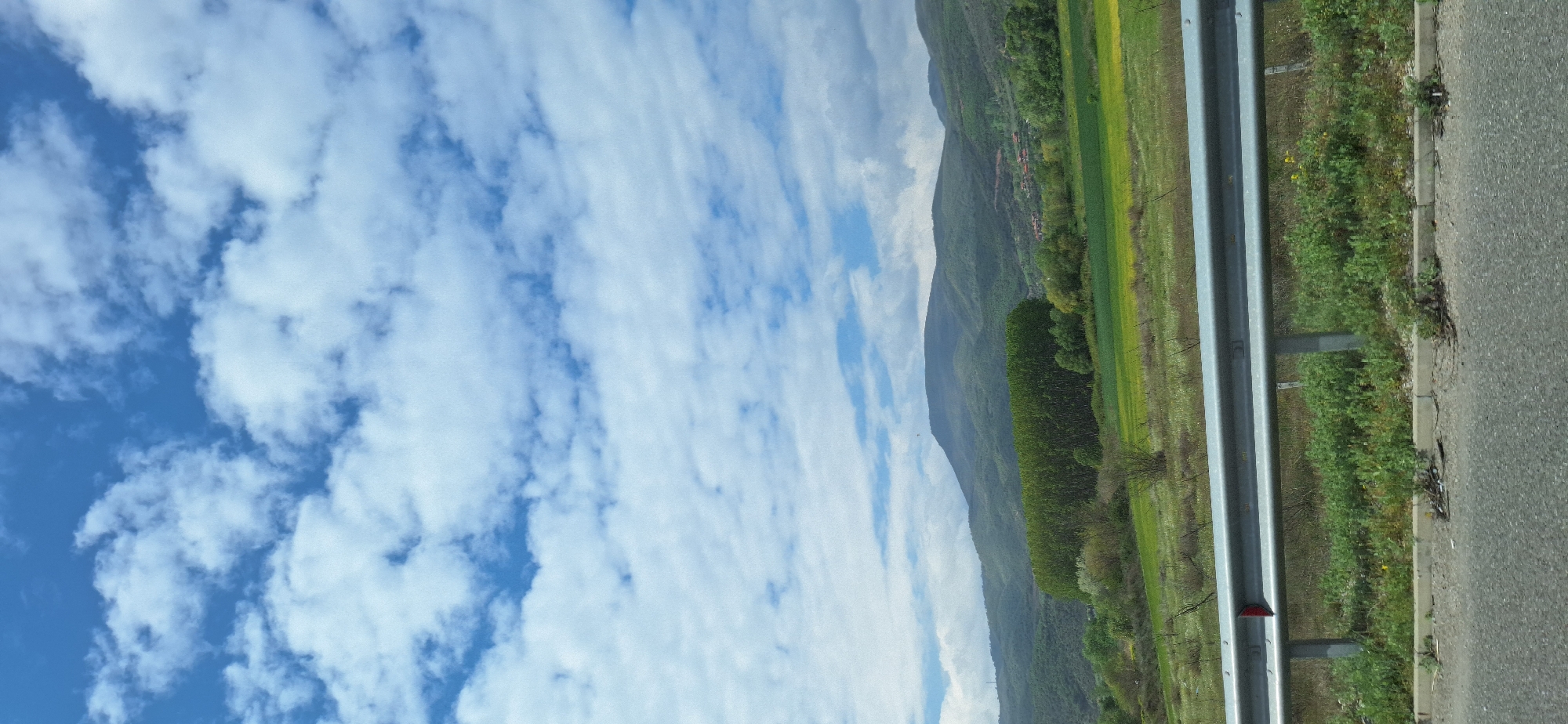A landscape photograph taken from a moving vehicle depicts a blue sky dotted with fluffy white clouds filling the left side of the frame. The right side shows a roadside environment, including a metal guardrail, a grassy verge with some wildflowers, and a stretch of asphalt road. Beyond the roadside, a green field extends towards a mountainous terrain. The location is Valkovo, Bulgaria, based on the landscape and vegetation. There is no visible human activity.
