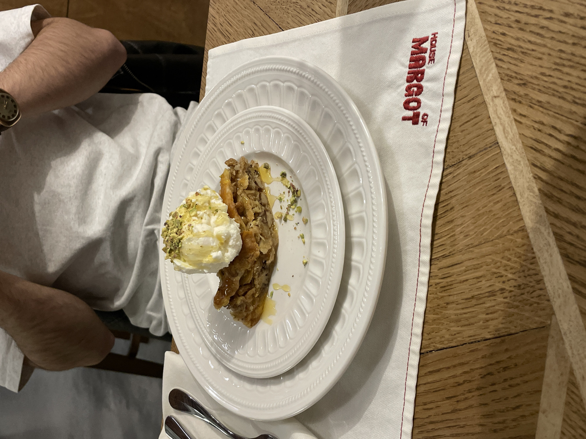 Here is a detailed description of the image:
A high-angle shot shows a plate of food on a white napkin with red trim and the embroidered words "HOUSE OF MARGOT" in red. The food appears to be baklava served with a dollop of whipped cream and chopped pistachios. The plate is white with a decorative, scalloped edge. Two silver utensils (likely a fork and spoon) are visible below the plate.
A person wearing a white shirt and a watch is partially visible in the upper-left corner of the frame. The table beneath the napkin is made of light-colored wood.
Based on the name "HOUSE OF MARGOT," the food is likely being served at the House of Margot restaurant, located in Yerevan, Armenia.