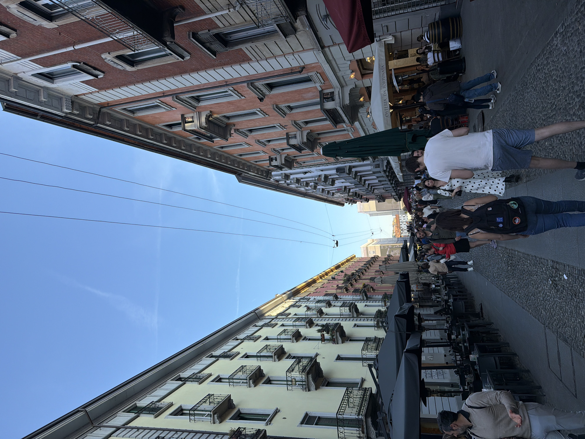 This outdoor shot depicts a street scene in Milan, Italy. The perspective is from ground level, looking up at the buildings lining the street. On the left side of the frame, a building with yellow facades and black wrought-iron balconies rises towards the sky. On the right side, a building with orange facades and similar balconies complements the scene. Overhead, power lines span the gap between the buildings, converging towards the vanishing point in the distance.
A bustling pedestrian street is visible between the buildings. People of various ages and attire are walking and interacting. Some individuals are looking at shop windows or engaging in conversations. Outdoor seating areas with tables and chairs are set up along the right side of the street, with some patrons seated and enjoying refreshments.
The sky above is a clear, gradient blue, contrasting with the urban environment. The overall atmosphere suggests a vibrant and lively urban scene in Milan.