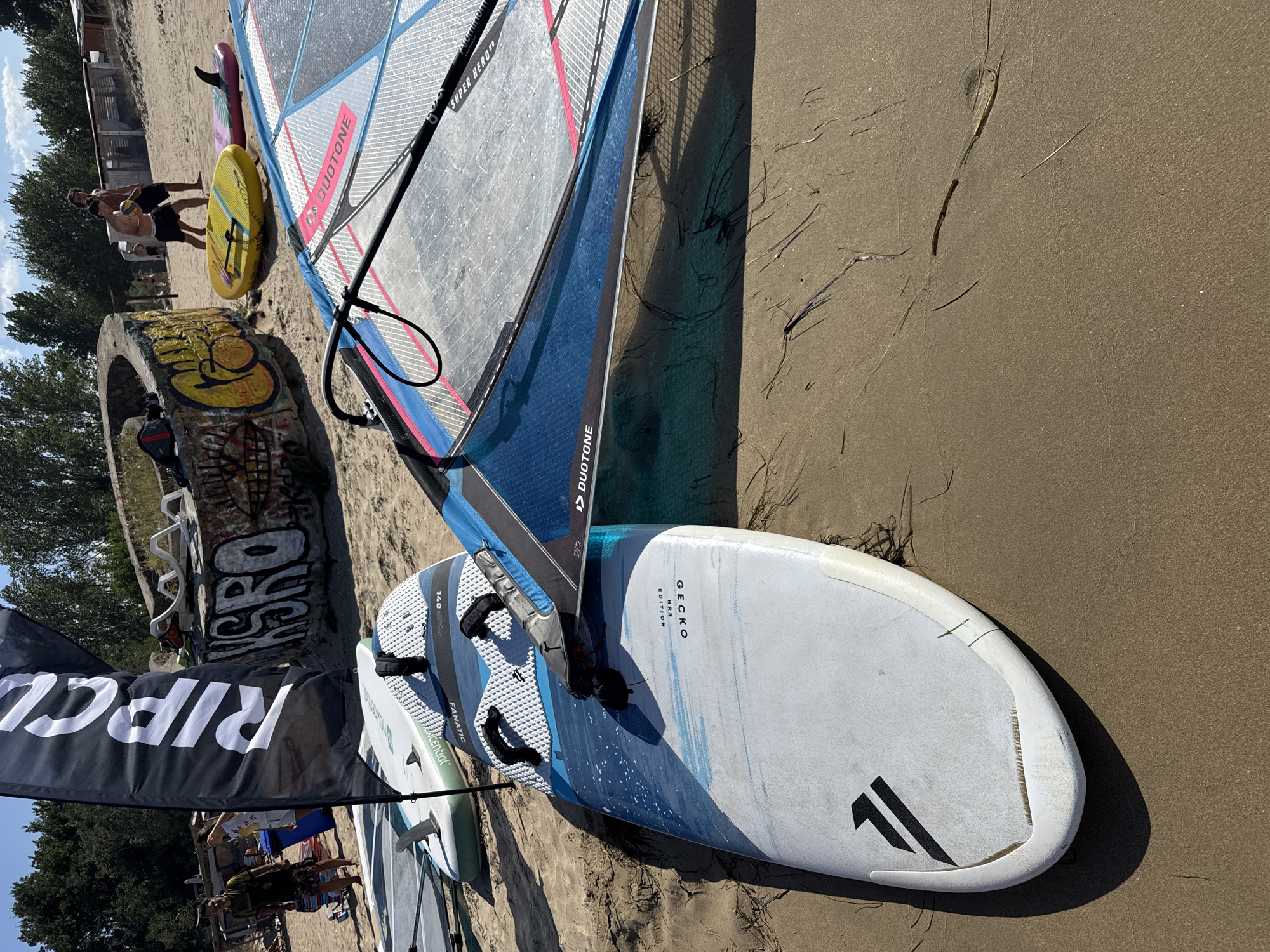 The image captures a windsailing scene on a sandy beach, likely in Sozopol, Bulgaria, given the context of previous images. In the foreground, a windsurfing sail, predominantly blue and white with pink accents, is laid out on the sand. The sail bears the brand name "DUOTONE" and "SUPER HERO". Two windsurf boards are positioned in front of the sail. The board closer to the viewer is white with blue abstract markings and the brand name "FANATIC" and "GECKO HRS EDITION" are visible. The second board, partially obscured, is white and green with a fin.
In the background, a black flag with white lettering that reads "RIPOLL" is planted in the sand. Further back, on the beach, several people are visible. Two shirtless men, one holding a yellow ball, are standing near a graffiti-covered concrete structure. Other individuals can be seen further down the beach, near some beach structures. The environment is a sun-drenched beach with a clear sky and trees bordering the background. The sand appears dry and speckled with dried seaweed.