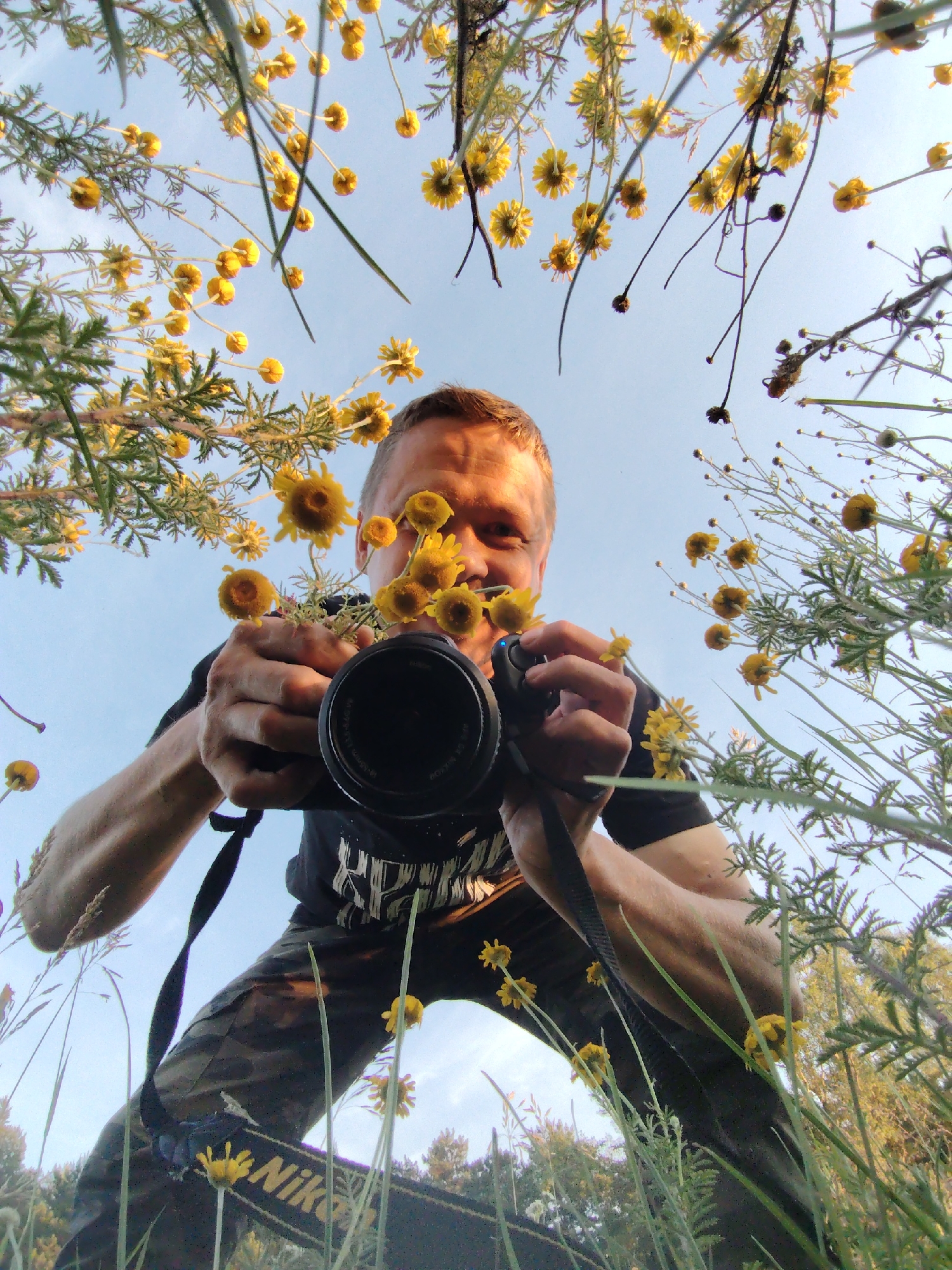 A low-angle, wide shot captures a man holding a camera amidst yellow wildflowers and green foliage under a clear sky, possibly in Chaikovsky, Russia. The man, facing the camera, has short hair and is wearing a black t-shirt with a graphic print and camouflage pants. He is holding a black camera with a lens that is in focus, positioned to take a photo of the sky and surrounding flowers. His right hand is on the camera, while his left hand is also near the camera, possibly adjusting settings. The yellow flowers are abundant, with some stems and leaves arching over the man and the camera. The background shows a bright, pale blue sky with hints of clouds. The lighting suggests it is daytime, possibly late afternoon or early morning given the soft light. The composition creates a sense of immersion within the natural environment.