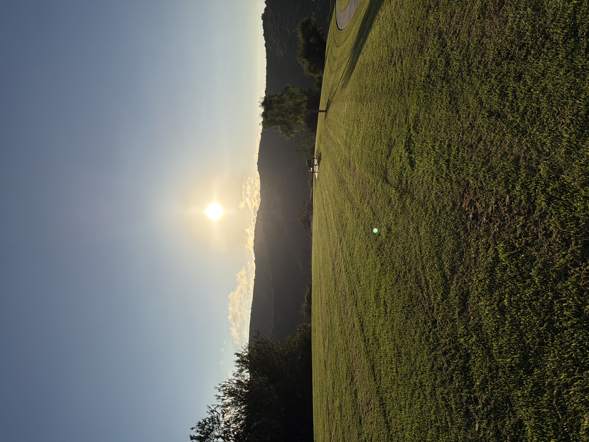 This is a photograph of a sunlit grassy knoll with a walking path and trees in the background. The sun is visible in the upper right portion of the frame, casting a warm glow over the landscape. In the middle ground, there is a park bench situated on the knoll. A lone tree with sparse foliage stands to the right of the bench. A paved walking path curves along the edge of the knoll in the right side of the image. The background consists of rolling hills covered in vegetation. The sky is clear and blue with a few wispy clouds. The image appears to be taken in Aliso Viejo, California, USA, given the characteristic rolling hills and coastal vegetation.