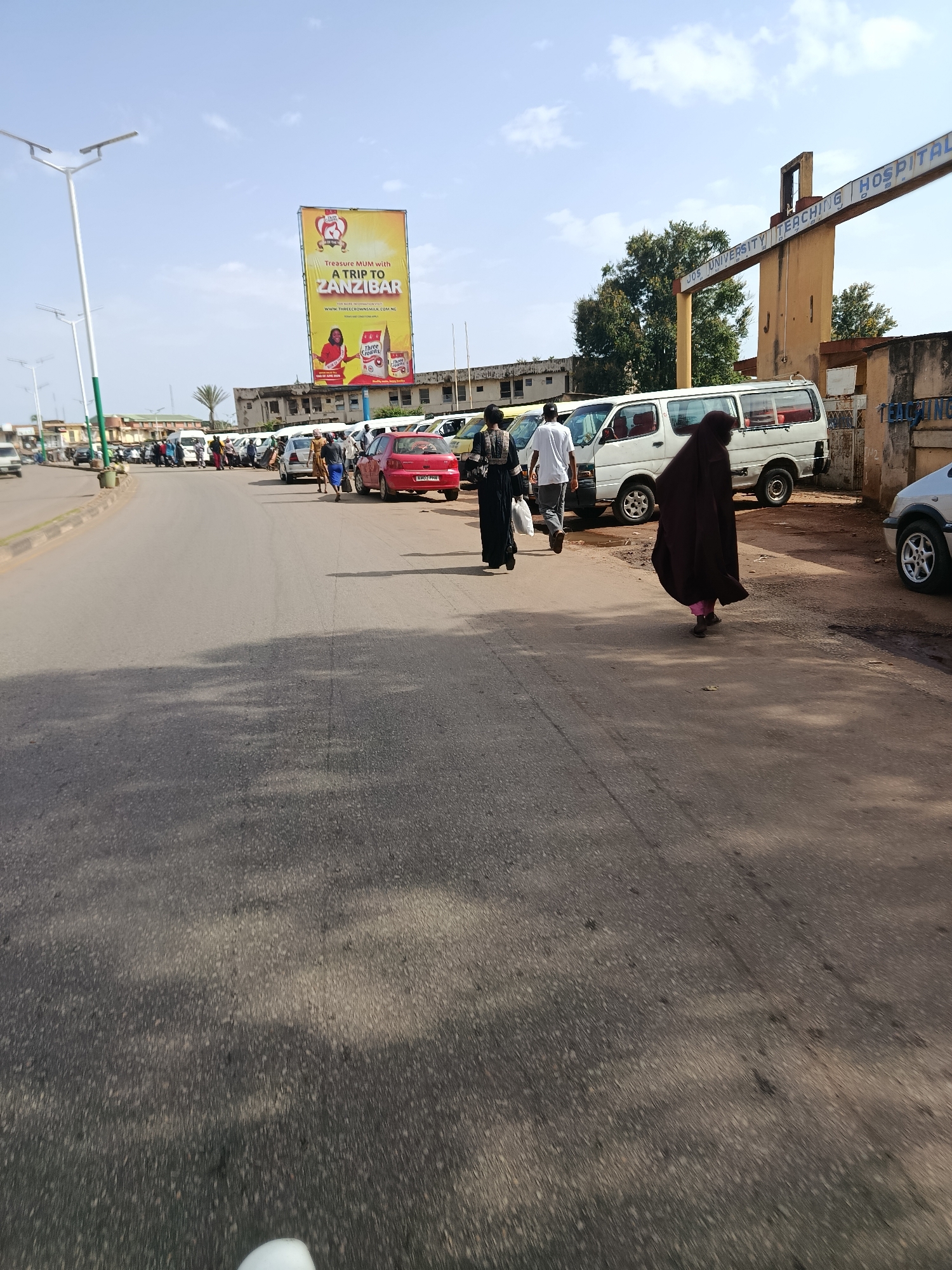 The image captures a street scene in Jos, Nigeria, on a clear, sunny day. The focus is on a street lined with parked vehicles, primarily white minivans, and a few cars. Several people are walking along the street and sidewalks, some appearing to be waiting for transportation.
In the foreground, the asphalt road is visible, with shadows from unseen objects falling across it. Further down the street, a red car is parked, and several individuals are walking in the same direction.
To the right of the road, a large billboard displays an advertisement for "Three Crowns Milk" with the text "Treasure Mum with a trip to Zanzibar." Below this, another line reads "www.threecrownsmilk.com.ng." Adjacent to the billboard is the entrance to "JOS UNIVERSITY TEACHING HOSPITAL," indicated by text on an overhead archway.
A woman in a dark hijab and pink pants walks away from the camera towards the hospital entrance, while another person in a black abaya and headscarf walks in the opposite direction. A man in a white t-shirt and dark pants is also visible, walking with the woman in the abaya.
The overall impression is a busy street scene, likely in the morning or afternoon given the bright sunlight and clear skies, with people going about their daily activities near a significant public institution.