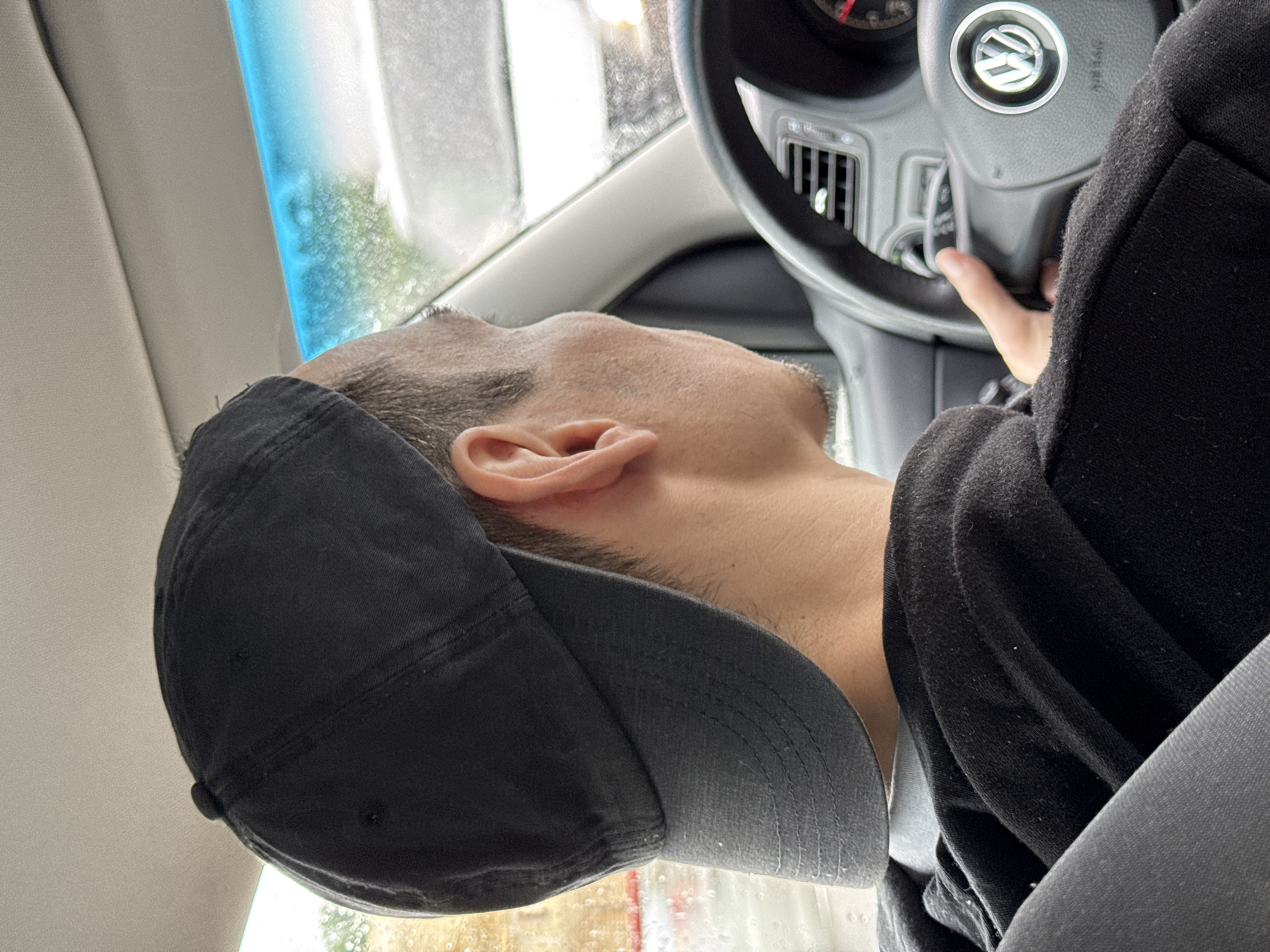 The image depicts a man in the driver's seat of a car, wearing a black cap and a black hoodie. His face is visible, and he appears to be looking at the steering wheel which has a Volkswagen logo. His hand rests on the steering wheel. A seatbelt is visible across his torso. Rain can be seen on the windshield. The location is not identifiable from this image.