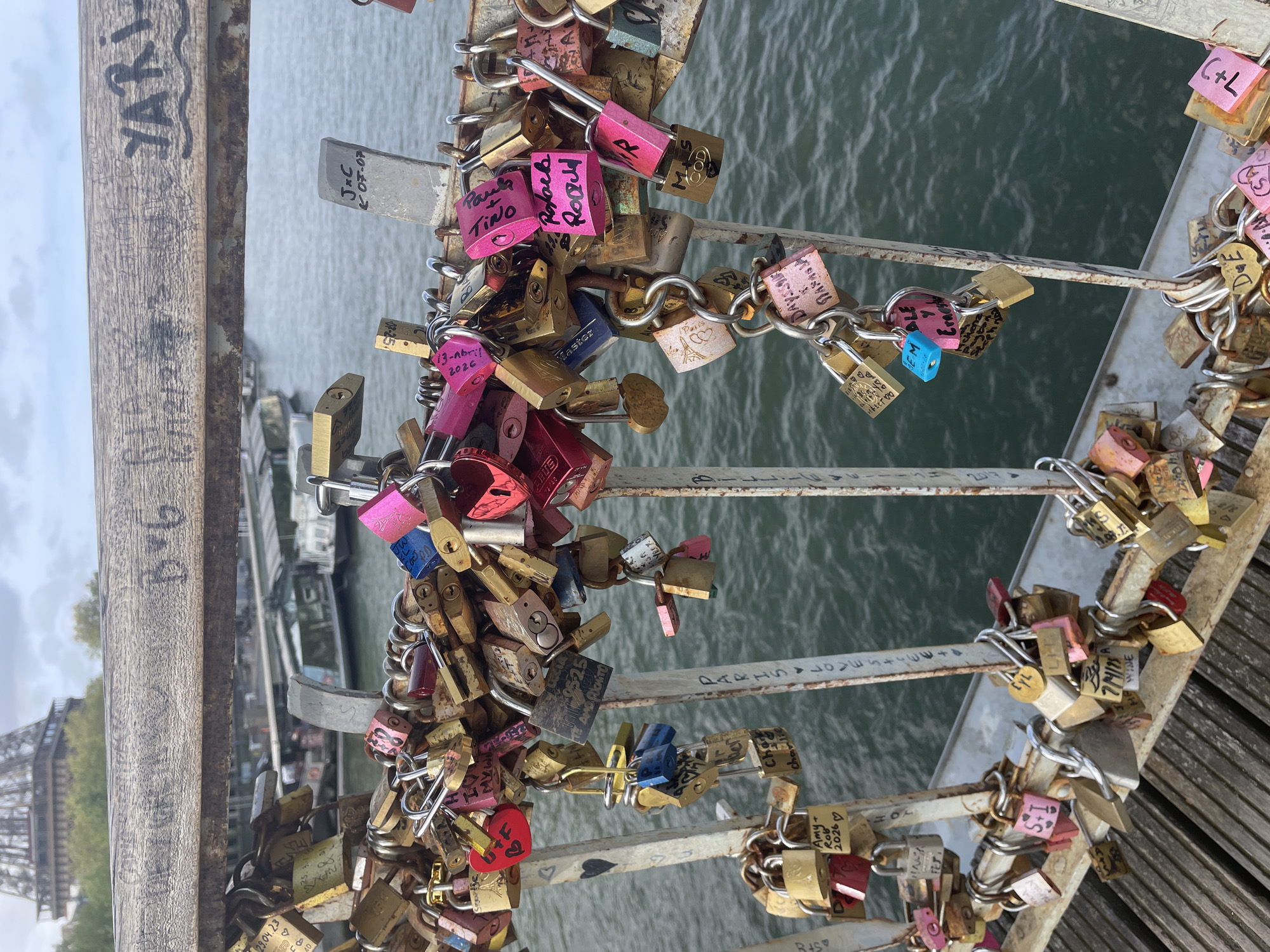 The image depicts a section of a bridge in Paris, France, heavily adorned with "love locks." These padlocks, attached by couples to symbolize their affection, cluster densely on the bridge's railings and chains, with inscriptions in various languages including names, initials, dates, and personal messages. The locks are of different colors, sizes, and materials, some connected by chains. Below the railings, the river Seine can be seen. In the background, the iconic Eiffel Tower is partially visible. Graffiti, including inscriptions and dates such as "YARI," is present on the bridge's wooden framework.