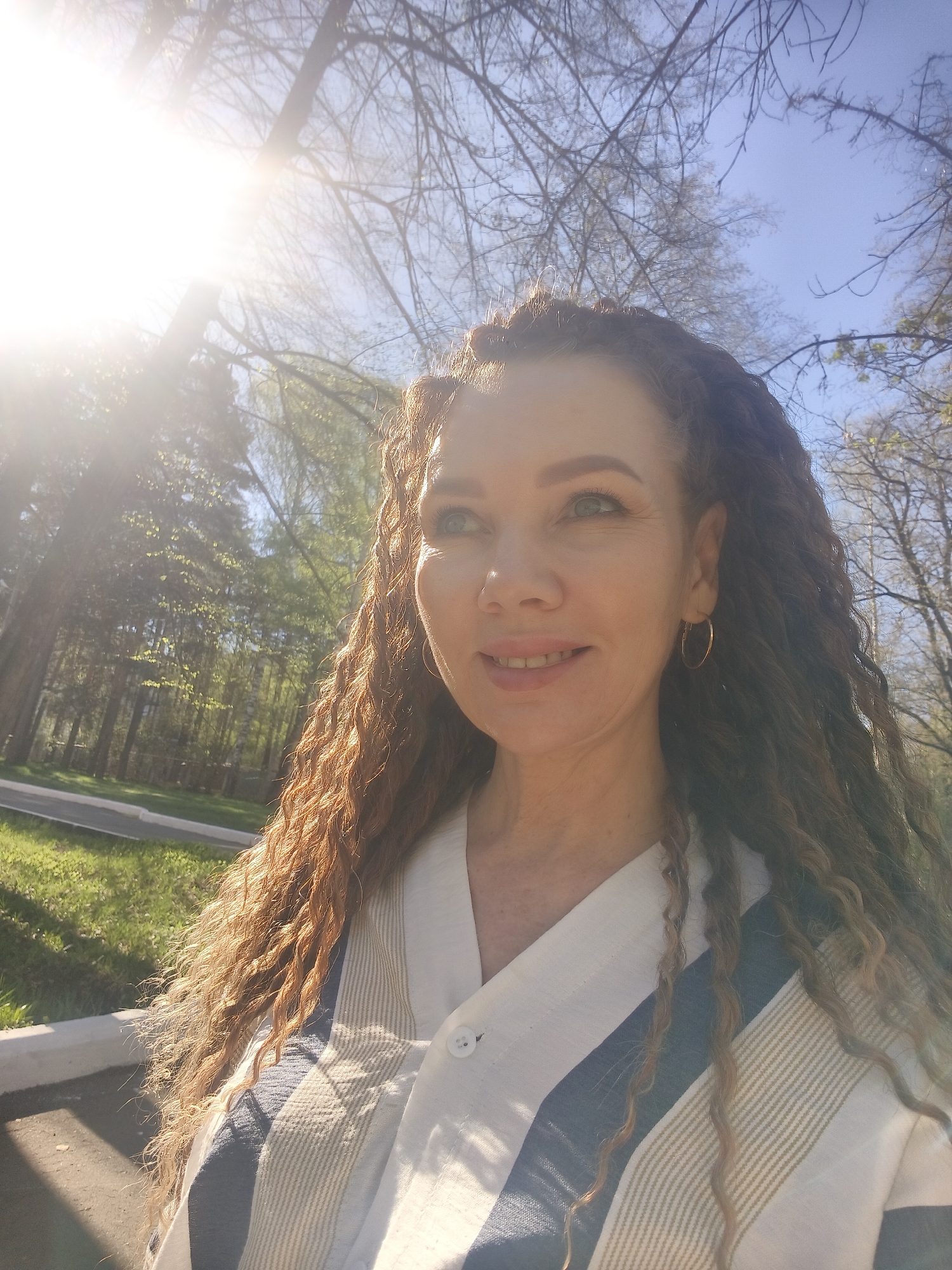A woman with curly brown hair is outdoors in a park or wooded area. She is wearing gold hoop earrings and a striped button-down shirt with white, tan, and blue stripes. The location appears to be a city park or green space with trees and grass visible. The sun is bright, causing lens flare in the image. The exact city and location are unknown.