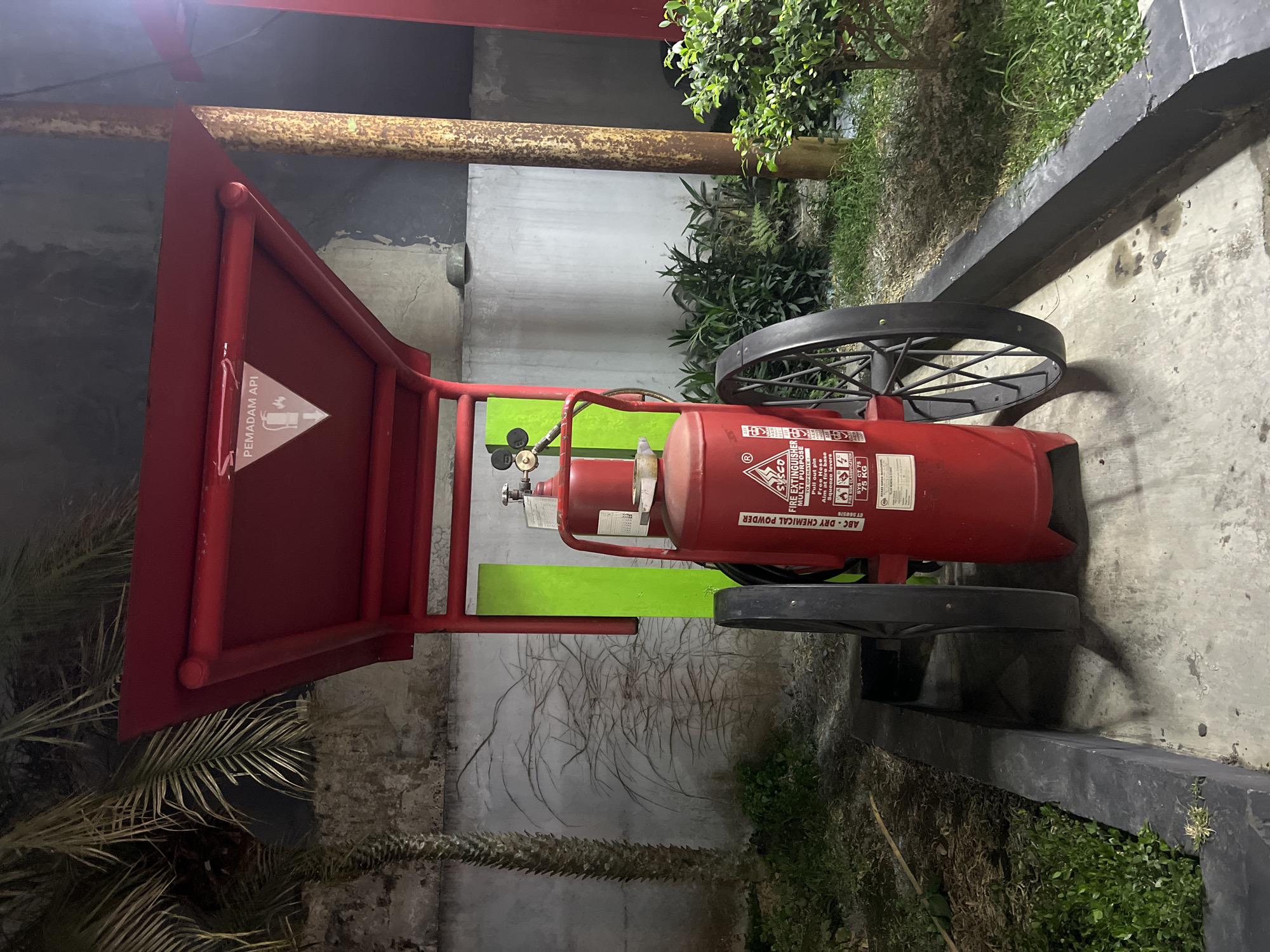 A large, red, wheeled fire extinguisher is positioned under a red canopy structure. The extinguisher has a white label with red lettering that reads "FIRE EXTINGUISHER MULTI PURPOSE ABC - DRY CHEMICAL POWDER". Above the fire extinguisher, on the underside of the canopy, a triangular white sign with a red border displays a symbol of a flame and the text "PEMADAM API". To the left of the extinguisher is a palm tree with green fronds. Behind the extinguisher is a gray concrete wall with abstract dark lines drawn on it, and a vertical bright green rectangular panel. To the right of the extinguisher is a rusty, cylindrical pole and a green bush. The ground in front of the extinguisher appears to be concrete, with a black curb to the left. The scene is outdoors at night, with artificial lighting illuminating the area. The image was captured in Magelang, Indonesia.