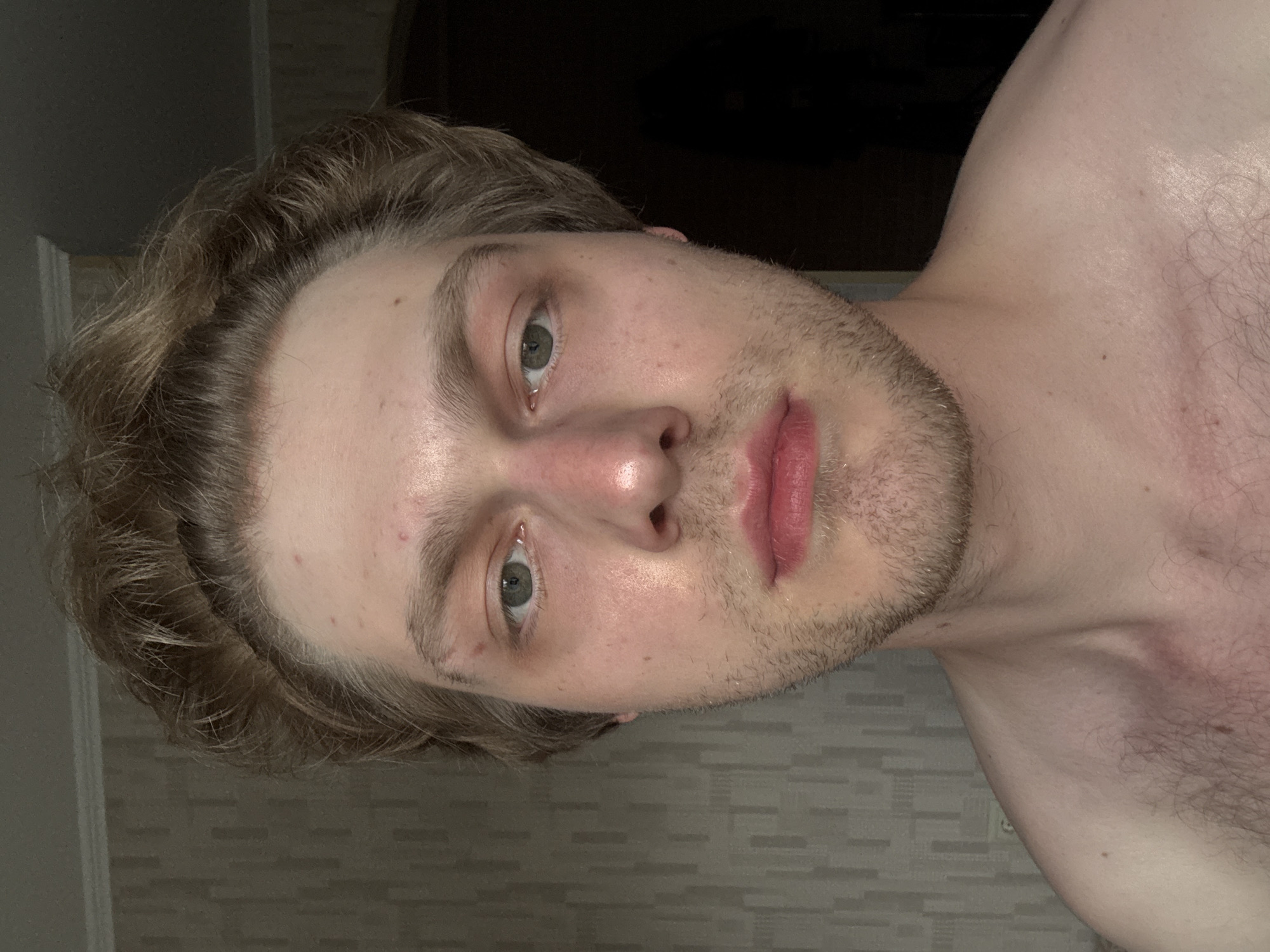 The media file is a close-up, vertically oriented, low-angle, selfie-style photograph of a young man's face and upper chest. He has light brown, wavy hair that is slightly tousled and falls around his forehead and temples. His skin is fair, with some redness on his cheeks and forehead, and a few small blemishes are visible. His eyes are a light green color, and his eyebrows are light brown. He has a light stubble on his jawline and upper lip. His lips are full and appear to be slightly glossy, with a pinkish-red hue. The background is dark, with some blurred elements that suggest an indoor setting with poor lighting. A textured wallpaper with a subtle brick or tile pattern is visible on the right side of the frame, partially obscured by the man's shoulder. The lighting is dim, creating strong shadows and highlighting the contours of his face. The overall mood is intimate and casual, possibly taken at night or in a dimly lit room. There is no visible text in the image.