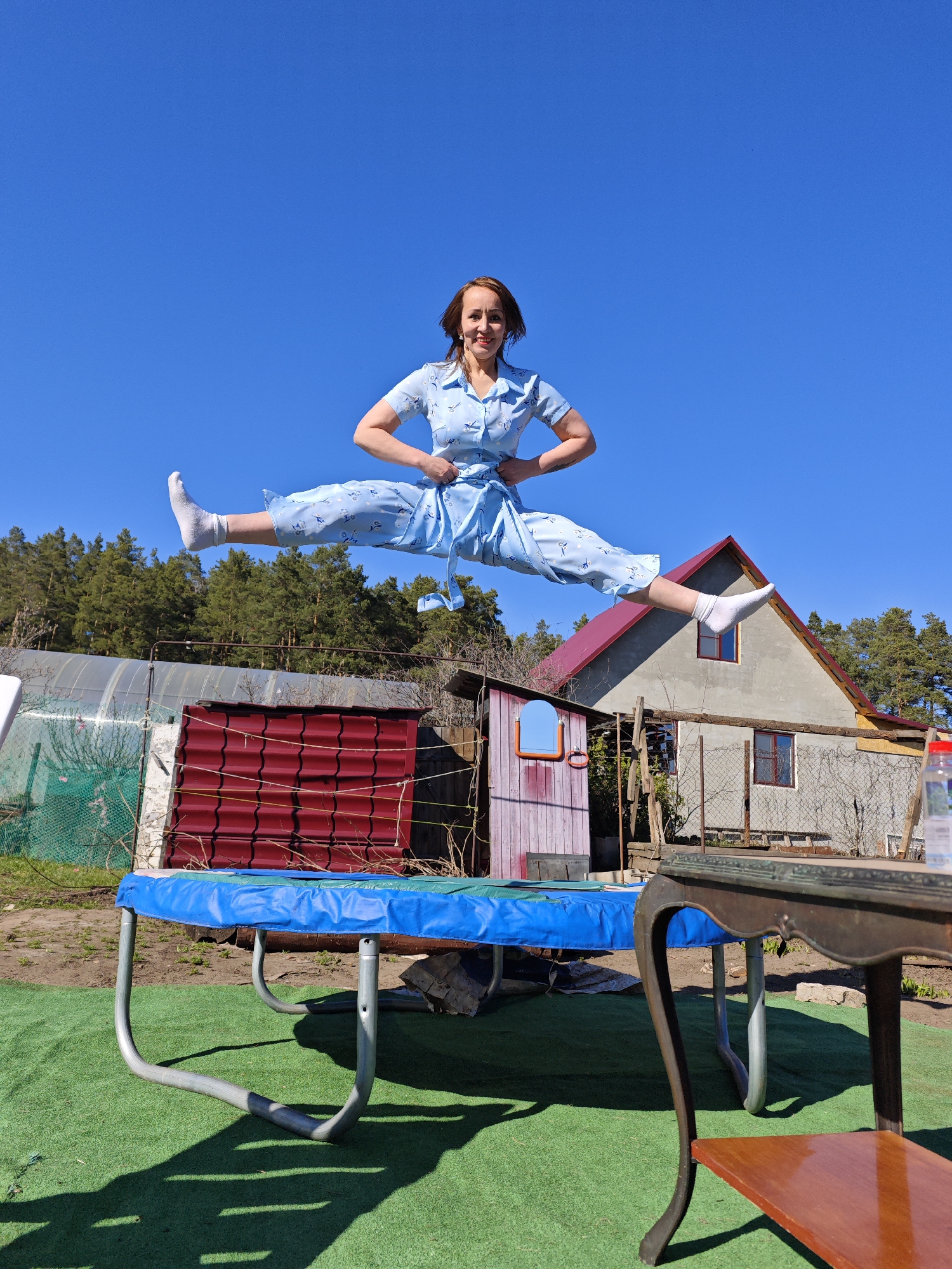 A woman is performing a split jump on a blue trampoline. She is wearing a light blue patterned button-down jumpsuit and white socks. The background includes a greenhouse, a shed with a red roof, a house with a red roof, and trees against a clear blue sky. A small table is visible in the lower right corner. The ground is covered in artificial green turf. Based on the style of buildings and the presence of a typical garden setup, this image could potentially be taken in a suburban or rural setting in Noviy, Russia.