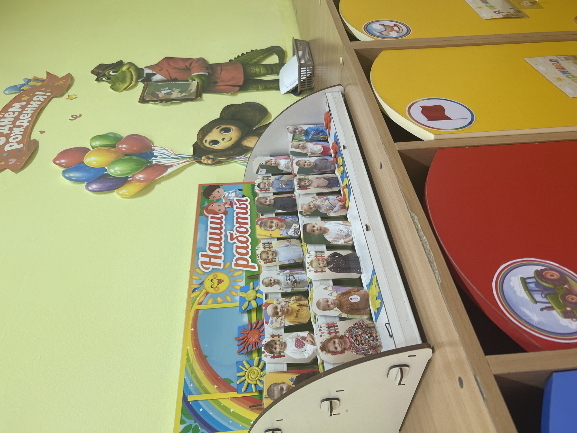 The image shows a colorful display on a wall, likely in a children's educational setting like a kindergarten or preschool in Gelendzhik, Russia. Dominating the left side of the frame is a whimsical cartoon alligator wearing a suit and hat, holding a framed picture. Next to it, a cartoon monkey with large eyes is depicted. Below these characters, a bunch of brightly colored balloons is shown, tied with strings.
A prominent display titled "Наши работы" (Our Work) is angled in the center of the image. This display features a rainbow arch and a smiling sun at the top. Below, there are multiple rectangular slots holding small portraits of children. These portraits appear to be of young children, some smiling and looking directly at the camera.
To the right, there are shelves containing various items. One shelf holds a yellow circular surface with a sticker of a rocking horse in a circular frame. Another shelf below it has a red circular surface with a sticker of a green tractor with a rainbow in the background. The overall impression is that of a vibrant and cheerful environment, designed for children. The lighting suggests it is daytime.