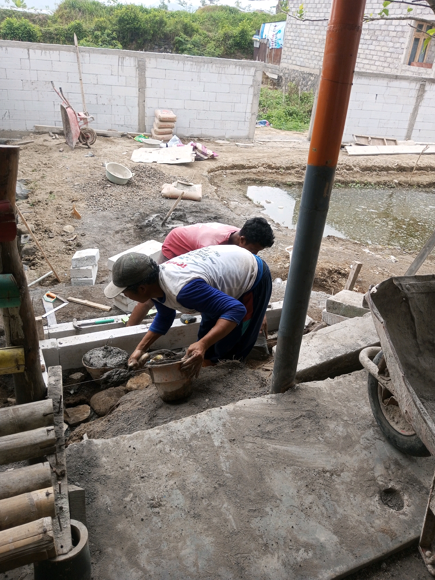 Two individuals, likely construction workers, are engaged in building work at a site in Kalibeber, Indonesia. The foreground is dominated by a concrete surface and the lower section of a bamboo structure. In the middle ground, one person wearing a white t-shirt with blue sleeves and a cap is kneeling and appears to be mixing or applying cement from a bucket. Another person, wearing a red shirt, is also present, partially visible and seemingly assisting. Construction materials such as cement bags, gravel, and what appear to be concrete blocks are scattered around the area. A wheelbarrow is visible on the right side of the image. The background features a partially constructed wall made of concrete blocks, greenery, and buildings. A body of water, possibly a pond, is situated behind the workers. The overall scene depicts an active construction site.