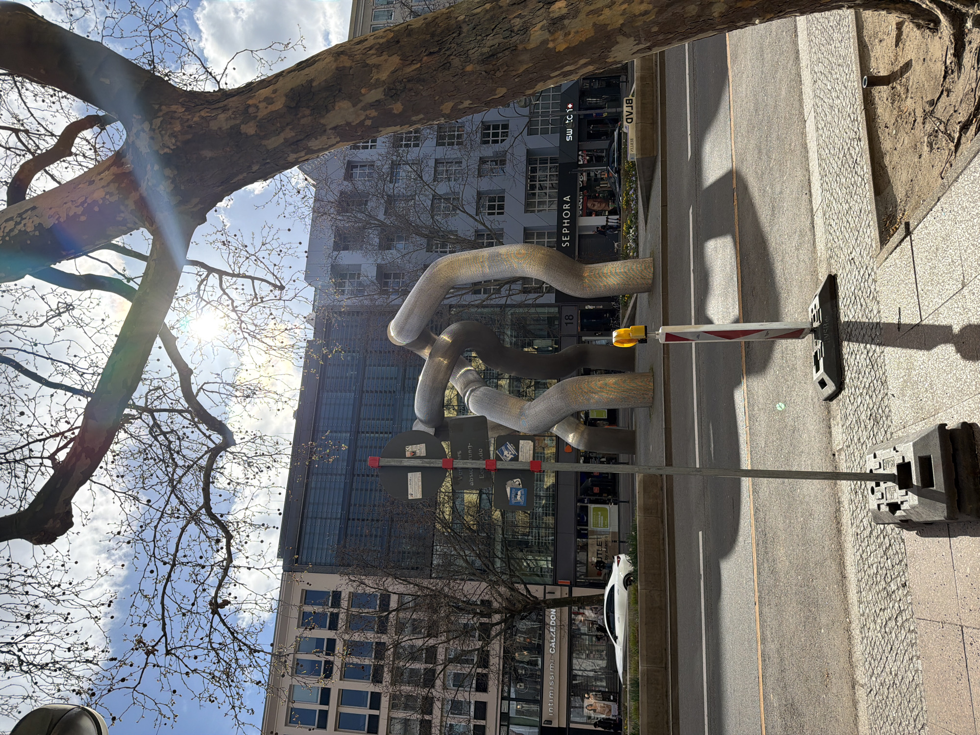 The image captures a street scene in Berlin, Germany. A prominent sculpture composed of interwoven, silver-colored tubular forms stands near the sidewalk. Behind it, there's a modern building with multiple windows, part of which reflects the sky. A tree with bare branches is positioned on the left side of the frame. On the ground level, businesses like "Sephora," "swatch+" and "intimissimi CALZEDONIA" are visible. A white car is parked alongside the street. A temporary traffic barrier with a yellow warning light is positioned at the sidewalk's edge.