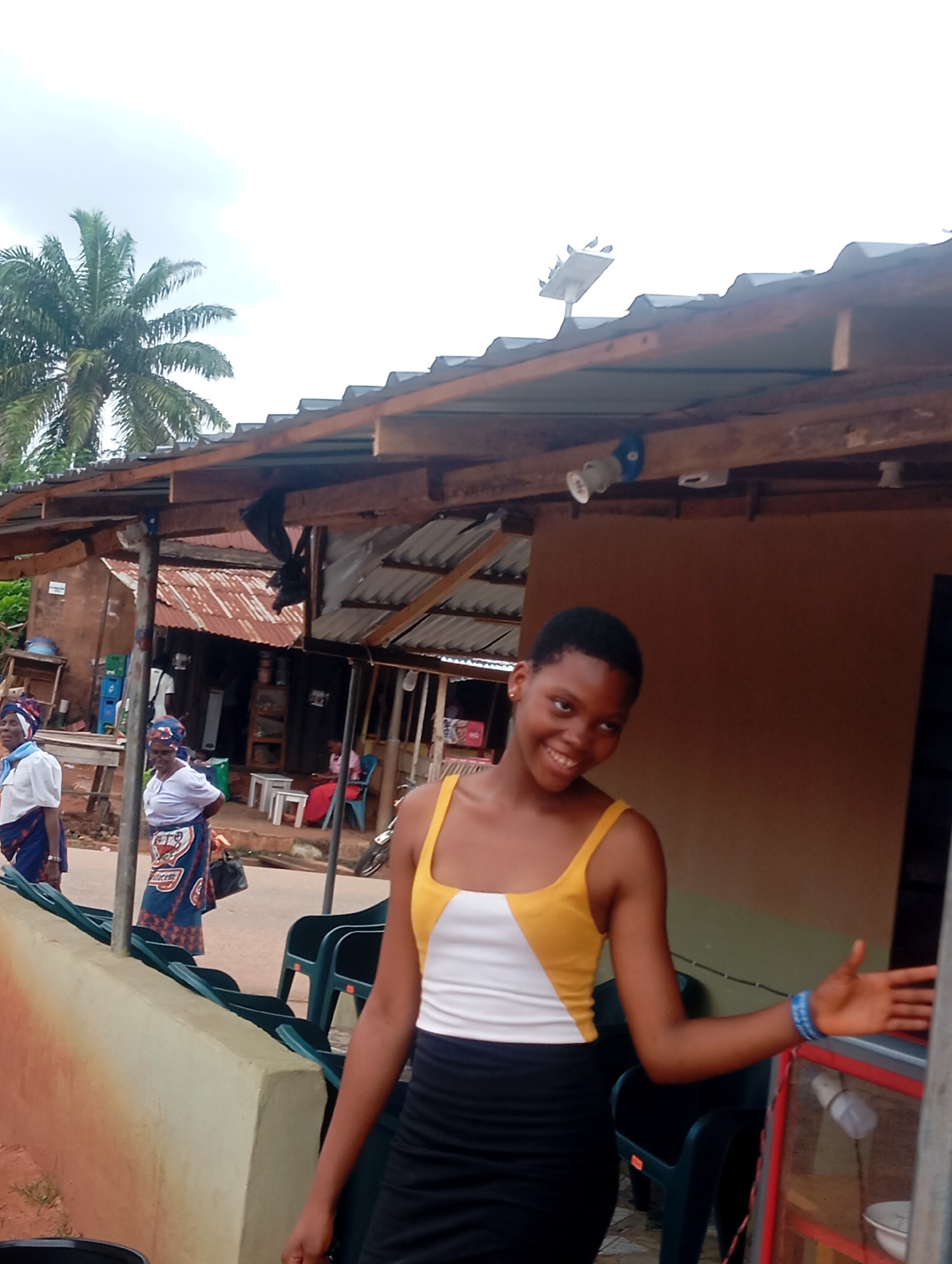 A young woman with short, dark hair and a bright smile stands in front of a covered market stall in Nsugbe, Nigeria. She wears a white and yellow sleeveless top and a black skirt. Her left hand is outstretched towards a glass display case, and her right arm is bent at the elbow. Behind her, two older women in traditional Nigerian attire, headwraps, and patterned wrappers are visible. One woman wears a light blue scarf and a white top, while the other has a blue and white patterned headwrap and a white blouse. The market stall is constructed with corrugated metal roofing and wooden beams. A palm tree is visible in the background. Several empty green plastic chairs are arranged in front of the stall, and a red and white cooler is to the right of the woman.