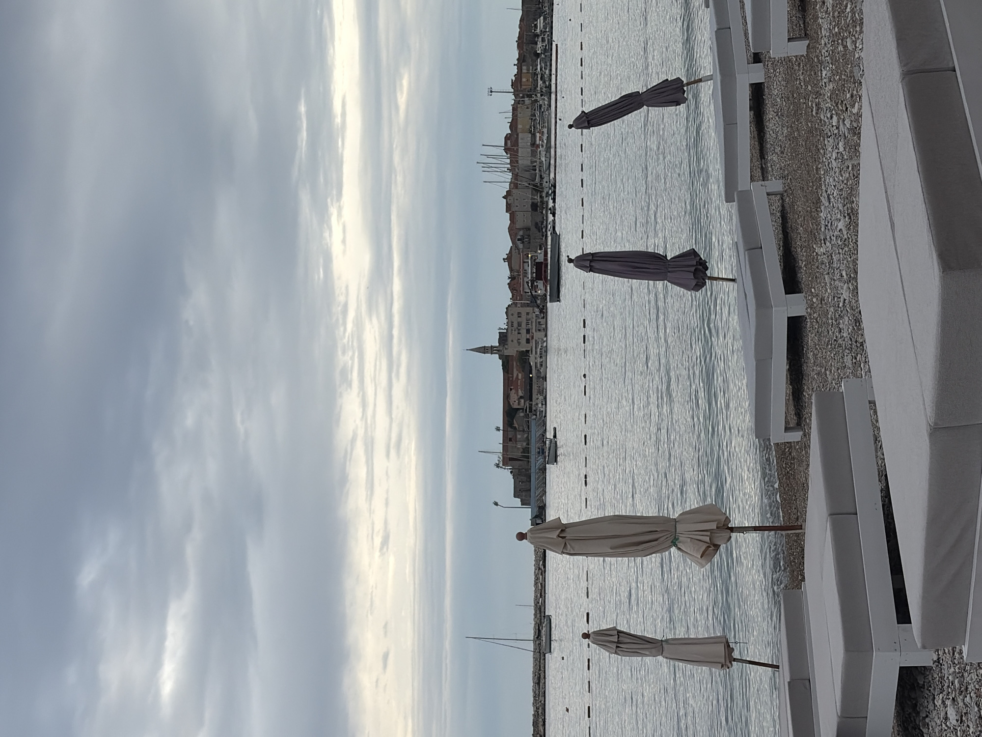 The image depicts a beach scene in Budva, Montenegro, on an overcast day. In the foreground, there are multiple rows of white beach chairs on a shingle beach. Closed beach umbrellas are interspersed among the chairs. The middle ground features the Adriatic Sea, with scattered sailboats and occasional ripples on the water's surface. In the background, the skyline of Budva is visible, characterized by buildings of varying heights and architectural styles. A church steeple stands out against the sky. The sky is heavily clouded with streaks of light peering through. The scene is quiet, with no apparent human activity visible.