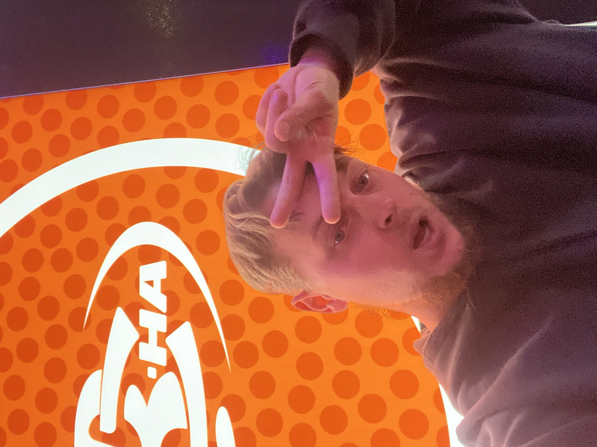 A man with blond hair and a red beard is posing in front of a Baskin-Robbins sign in Moscow, Russia. The man is wearing a dark-colored hoodie and is holding his hand up in a peace sign. The sign is orange with white lettering and polka dots. The lettering says "Baskin-Robbins".