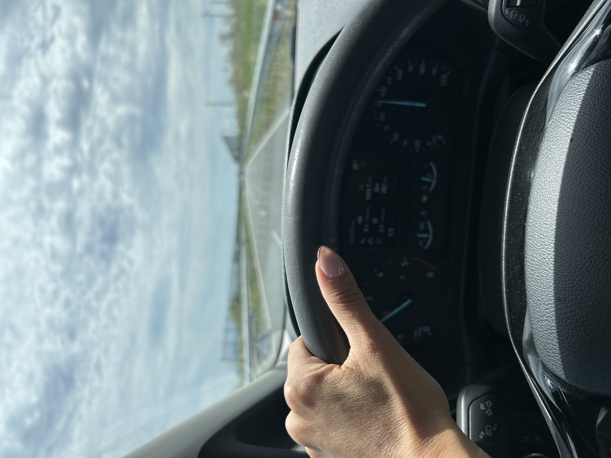 A first-person perspective shot of a person driving a car. The hand holding the steering wheel is in the foreground, and the dashboard with gauges and indicators is visible. The road ahead stretches into the distance, with a bright, cloudy sky above. The image appears to be taken from inside the car, looking forward. The location is stated to be the city of Aheloy, Bulgaria.