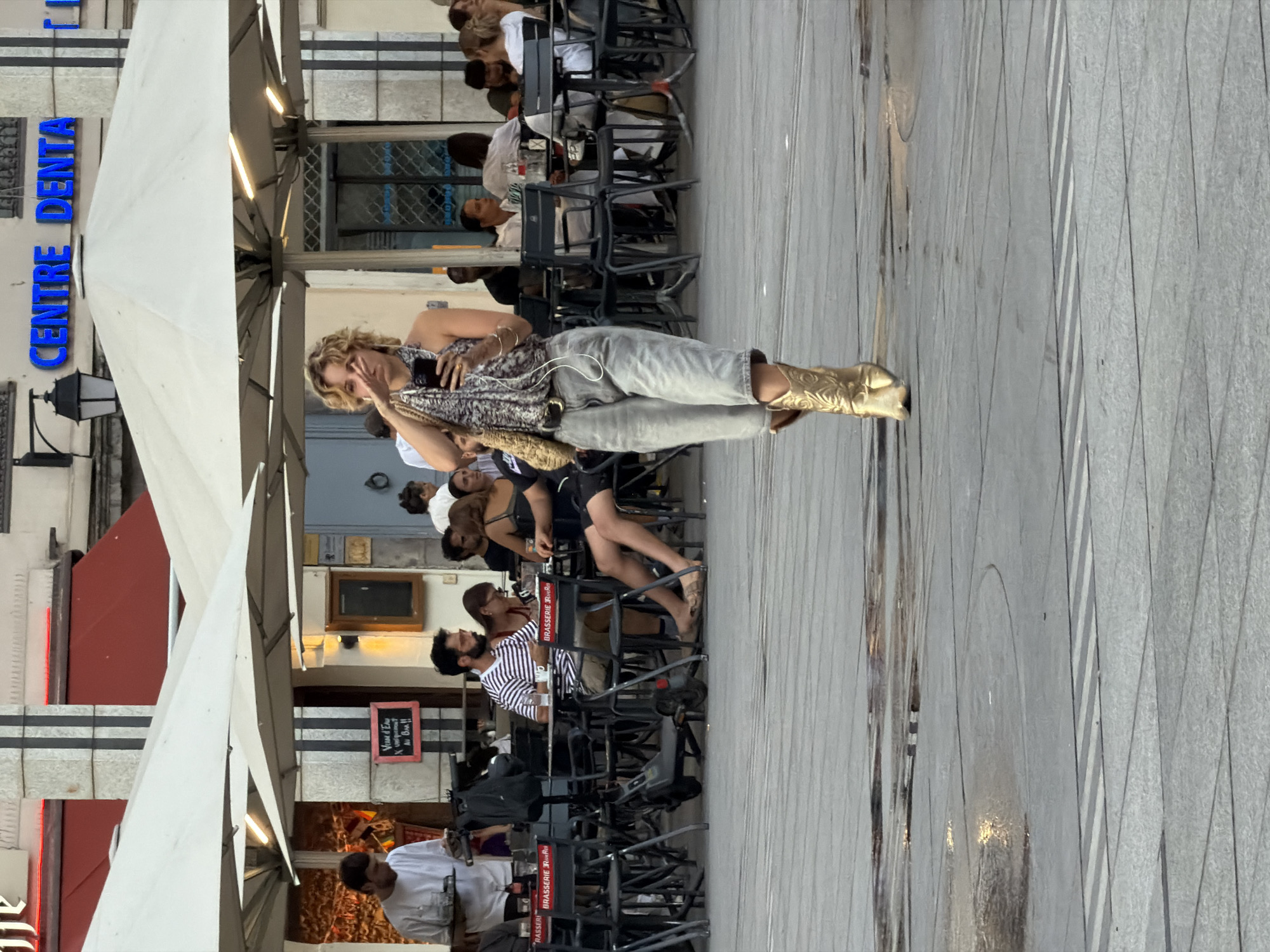 The image captures a street scene in Lyon, France, likely in an outdoor seating area of a cafe or restaurant. In the foreground, a woman walks towards the camera, holding a smartphone in her right hand and gesturing with her left. She is wearing a patterned halter top, loose-fitting grey pants, and striking gold cowboy boots. Behind her, patrons are seated at tables under large white umbrellas, some engaged in conversation or looking at their phones. The environment appears to be paved with stone tiles, with some wet patches reflecting the light, suggesting recent rain or cleaning. In the background, a building with blue signage that reads "CENTRE DENTA" is visible. An electric scooter is parked near a table with a man wearing a striped shirt. The overall atmosphere suggests a casual urban setting on a bright day.