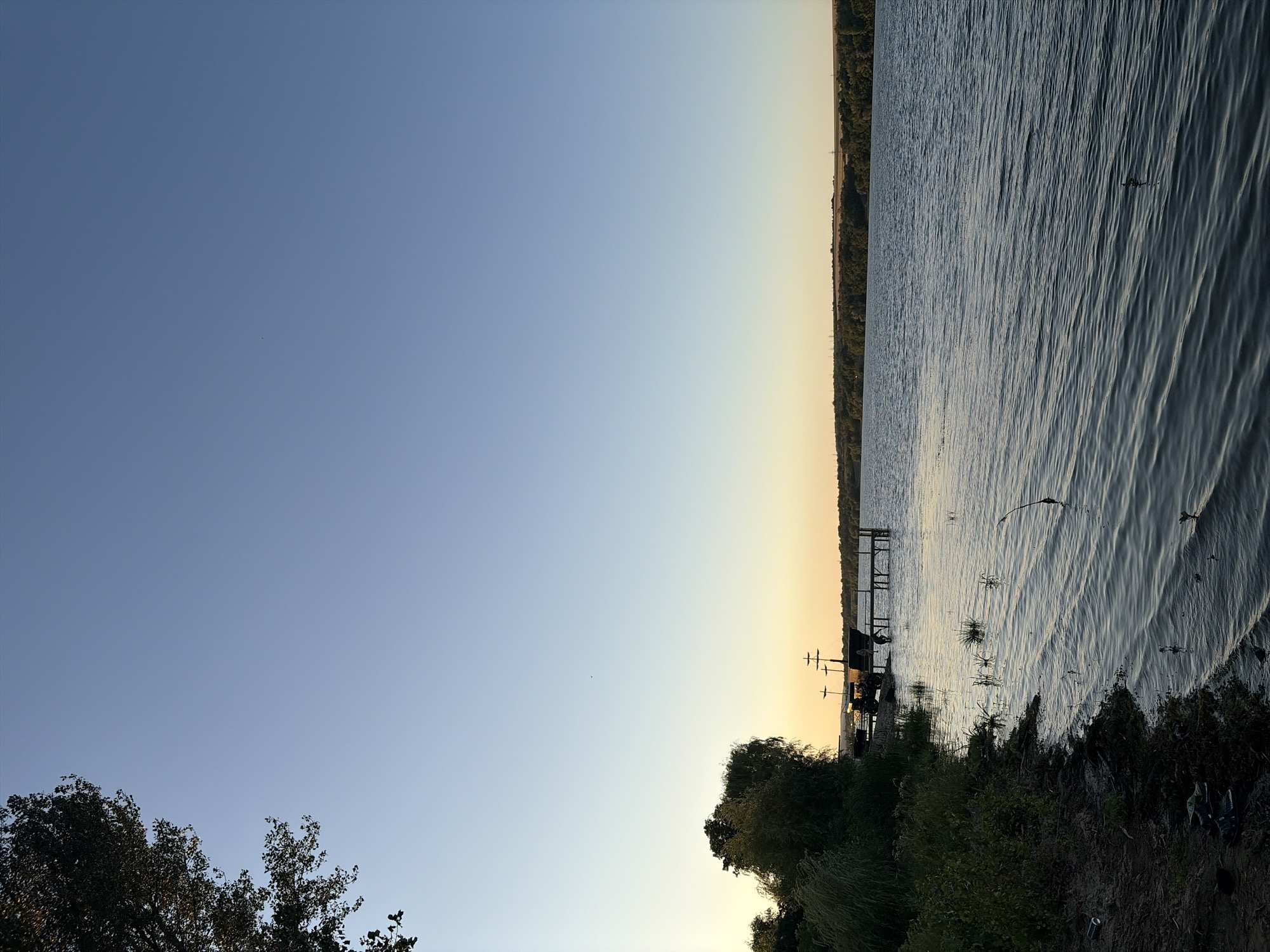 This is an outdoor, vertical, eye-level shot of a riverbank scene in Oxentea, Moldova, likely at dusk. The sky is a clear, pale blue with a faint orange glow near the horizon line. On the left side of the image, lush green foliage and trees line the shore, with a darker tree canopy extending into the upper left corner. The foreground consists of a sandy and grassy bank with a few discarded items including a blue can and a pair of flip-flops. A calm, dark blue river occupies the right two-thirds of the frame, its surface rippled by a gentle breeze. In the middle ground, a wooden dock extends into the river. A person is visible standing on the dock. On the dock, there are two boats, one of which appears to be a replica pirate ship with masts and sails. The distant shore is lined with a dense band of trees.
