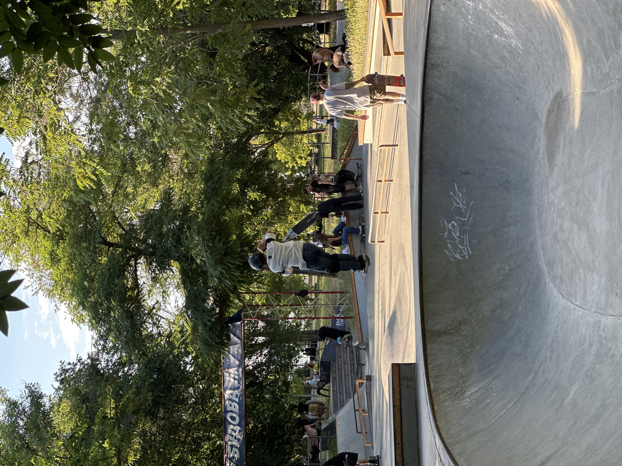The image depicts a lively outdoor skatepark scene during the day. In the foreground, a large concrete bowl with graffiti adorns its interior. In the mid-ground, a skateboarder in mid-air, wearing a helmet, elbow pads, and knee pads, is captured in action. Other individuals are present in the skatepark, some sitting on benches, others standing around, and a few are holding skateboards. In the background, a banner with text in a foreign language is visible, suspended above the skatepark structures. Lush green trees surround the area, suggesting a park or recreational space within the city. The lighting indicates a sunny day, with shadows cast on the concrete surfaces.