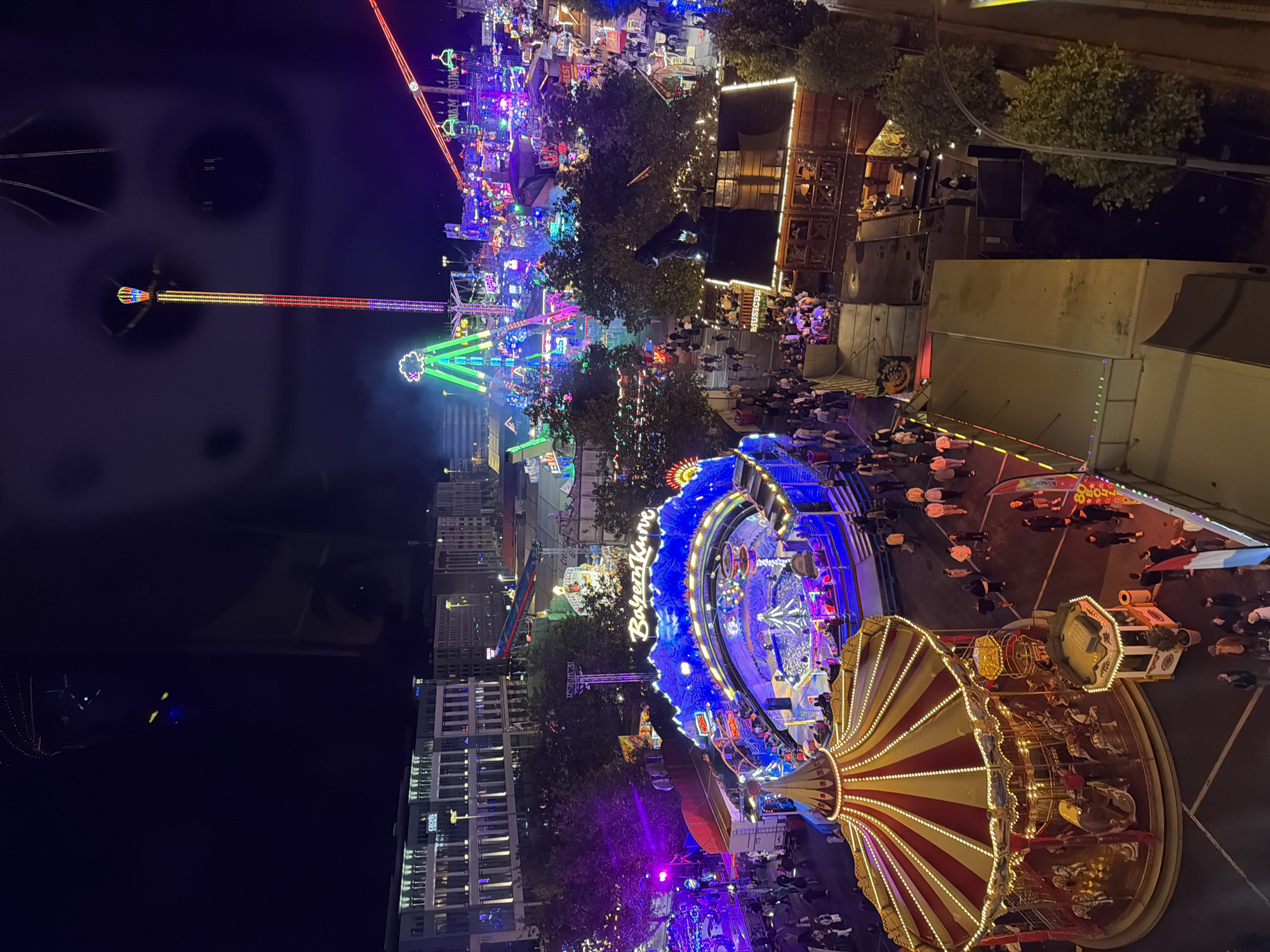 The image shows an aerial view of the Schueberfouer funfair in Luxembourg City at night. The funfair is illuminated with numerous colorful lights, highlighting various attractions. In the foreground, a carousel with a red and white striped roof is visible, with horses and other figures on display. To the right of the carousel, a small ticket booth with a potted plant is situated.
Further into the funfair, a large, brightly lit amusement ride with blue, illuminated mountain-like structures and a central area is prominent. People are seen walking around and enjoying the attractions. In the background, a tall, slender amusement ride with colorful lights, possibly a drop tower or similar structure, stands out against the dark sky. Several other rides and stalls, with a multitude of flashing lights in red, green, blue, and yellow, are scattered throughout the scene, creating a vibrant and festive atmosphere. Buildings with illuminated windows are visible in the far background, suggesting the funfair is located within an urban area. The overall scene depicts a bustling night at a large amusement park.