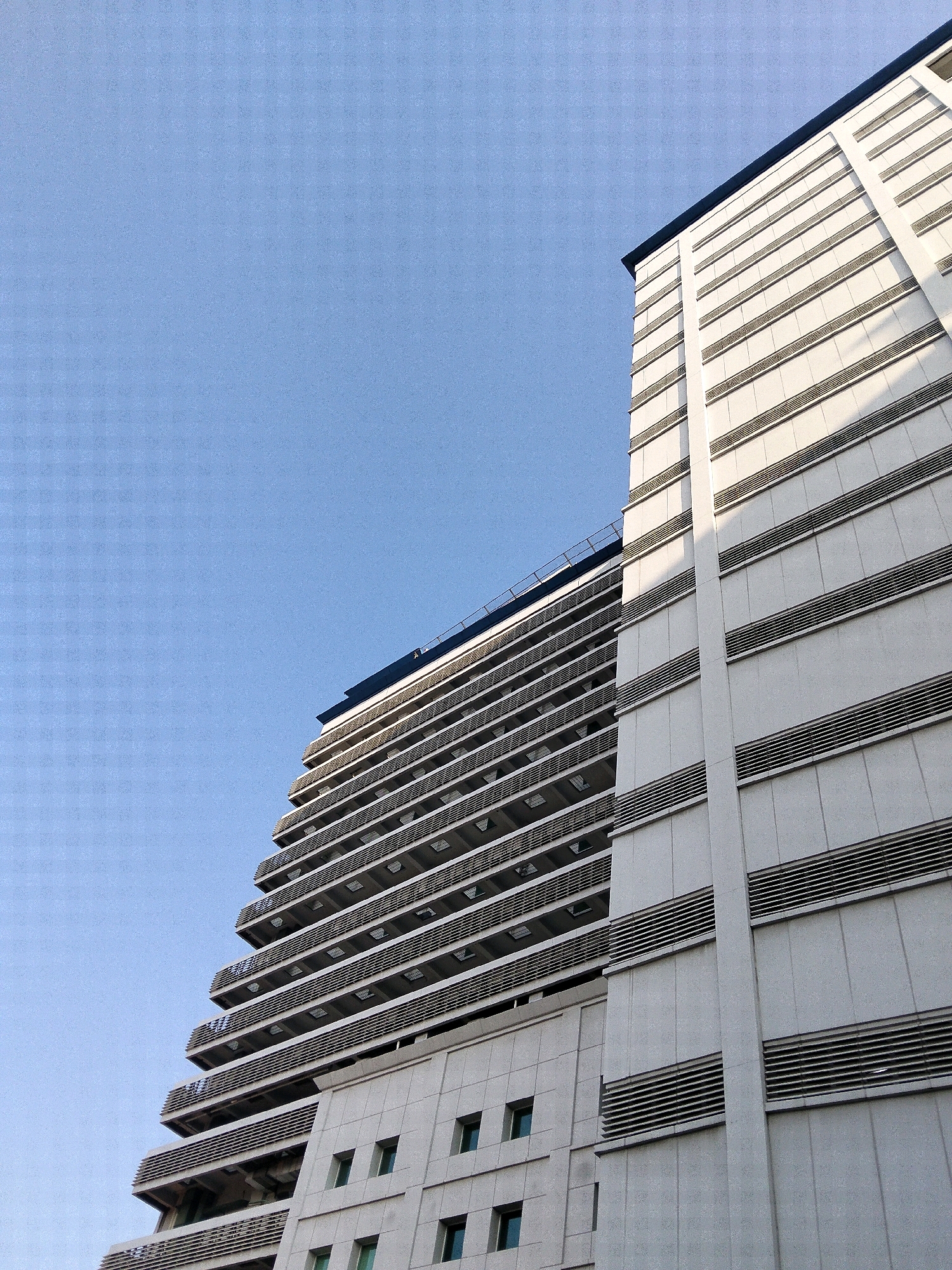 The image is an exterior, low-angle shot of two modern buildings against a clear blue sky. The building on the right is taller and more dominant, featuring a white facade with horizontal bands of dark-toned louvers or vents. It has a sharp, dark roofline. To the left, another building is visible, with a staggered, terraced design. This building also has a white facade and horizontal elements, with darker recessed areas suggesting balconies or windows. The overall scene depicts urban architecture under a bright, cloudless sky. The buildings appear to be located in Central Jakarta, Indonesia, based on external knowledge. There are no people or animals visible in the image.