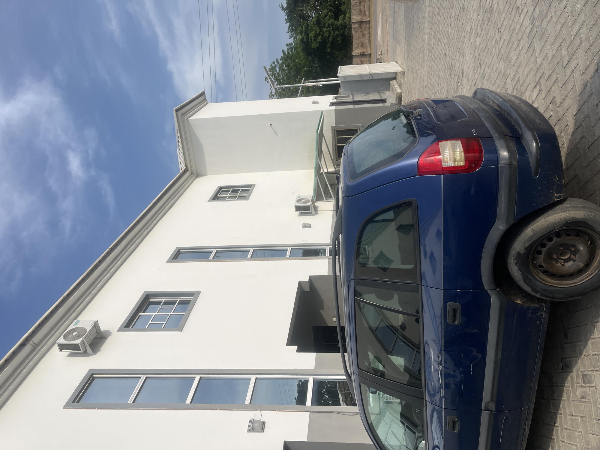 A blue minivan is parked on a paved driveway in front of a modern, two-story white building with gray accents. The building has several large, rectangular windows with gray frames. An outdoor air conditioning unit is mounted on the side of the building above a small, clear awning. The sky is blue with scattered white clouds. In the background, there are green trees and a low, concrete wall. The minivan shows signs of wear and tear, with visible scratches and dents on its body. The scene appears to be an exterior shot, likely in an urban or suburban setting.