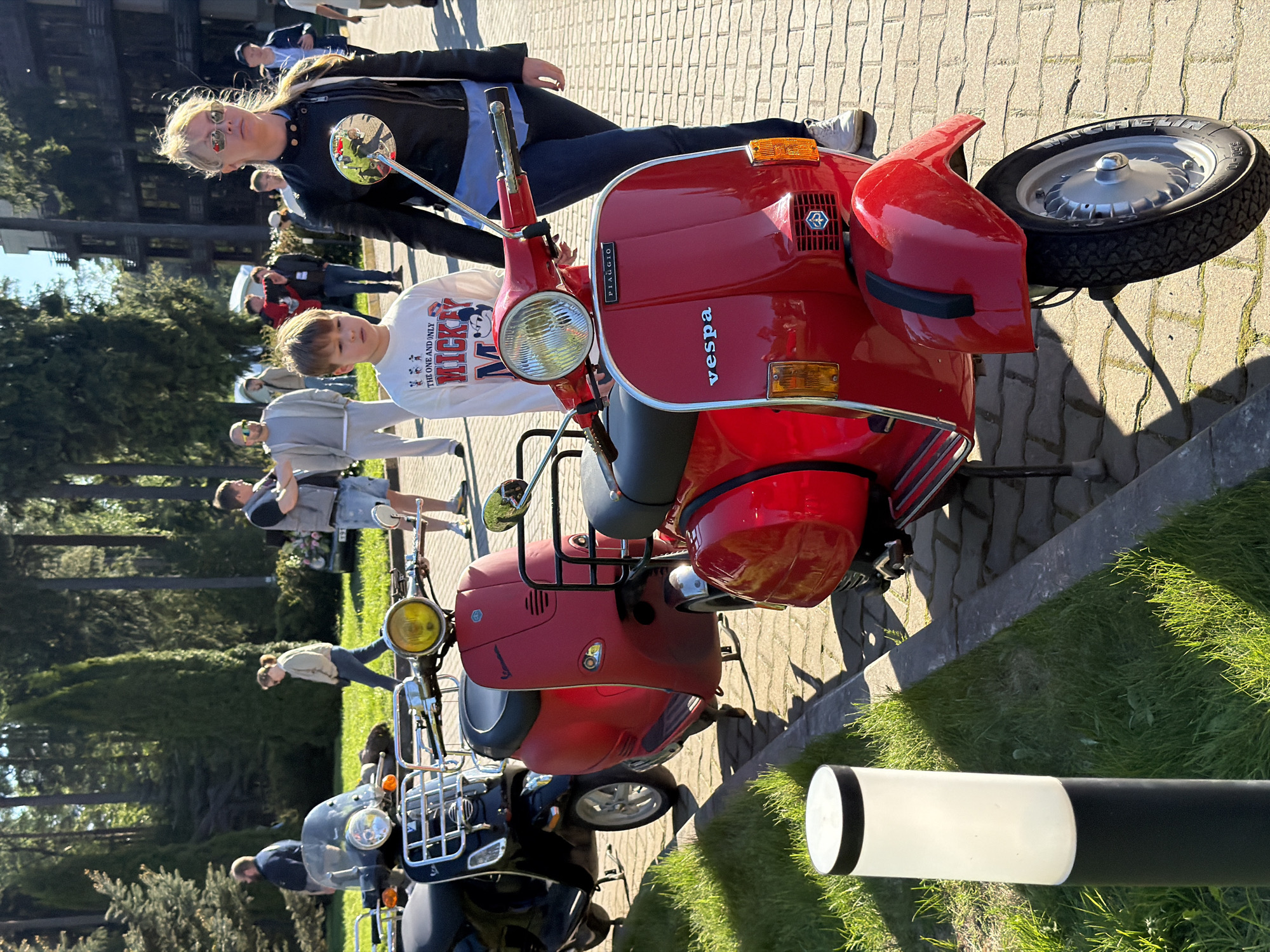 A red Vespa scooter is parked on a brick-paved surface in Repino, Russia, with a boy sitting on it and a woman leaning against it. There are more people and vehicles, including other scooters and cars, in the background. Trees are visible behind the crowd. A black and white pole stands near the scooters on a patch of grass. The day is sunny.