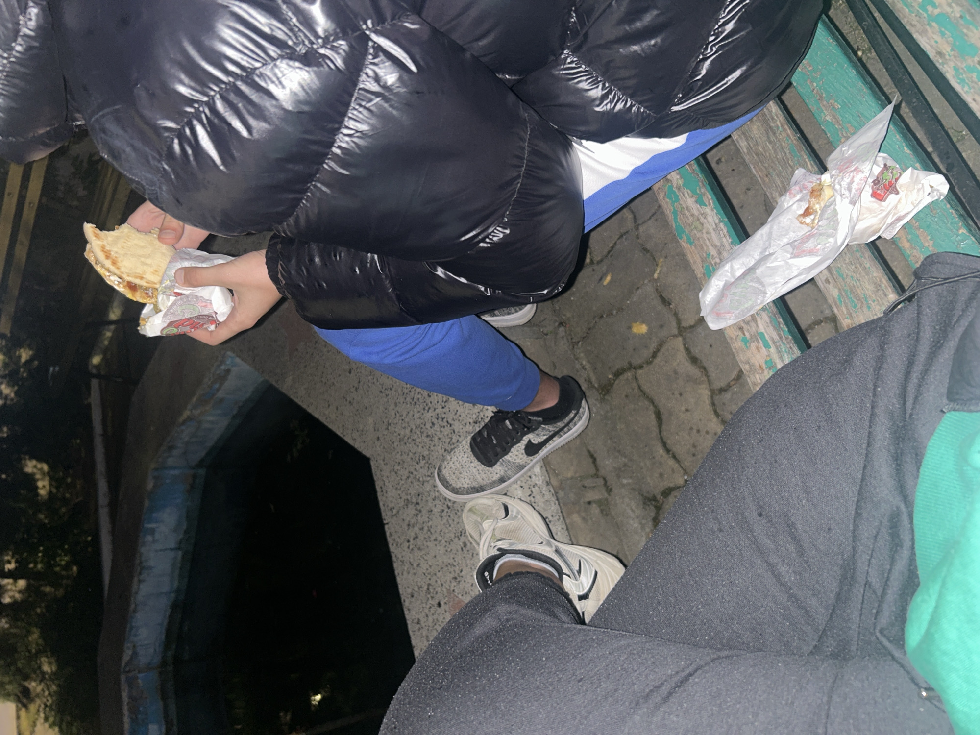 The image captures two individuals seated on a weathered, green-painted wooden bench alongside a waterway, possibly a canal or similar structure. The person on the left, wearing a black puffy jacket and blue pants, is eating a pita sandwich wrapped in paper. A crumpled piece of wrapper from a similar sandwich lies on the bench to their right. Their feet are adorned with gray and black Nike sneakers. The individual on the right is wearing dark gray sweatpants and white and beige sneakers, with a glimpse of a bright green garment underneath. The ground is paved with brick or concrete, and it appears to be nighttime. The city is unknown.