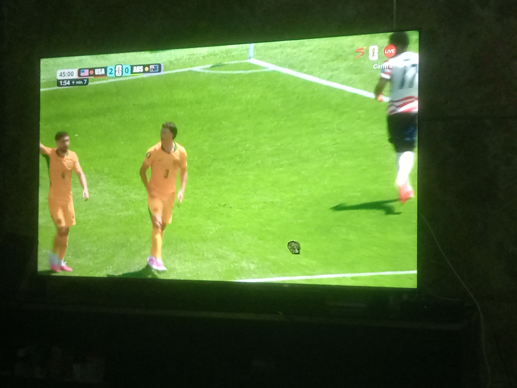 A television screen displays a soccer match. The score indicates USA 2, AUS 0. The time is 45:00, with 1:54 plus minutes added. Two soccer players in orange uniforms are on the green field. One player on the left is facing away from the camera and toward the right side of the screen. The second player, slightly to the right and further back, is also facing away from the camera and toward the right. A third player wearing a white and red striped jersey with the number 11 is visible in the background on the right side of the screen, running towards the left. There is a small dark object on the grass between the players. The image quality is poor, with significant glare and graininess.