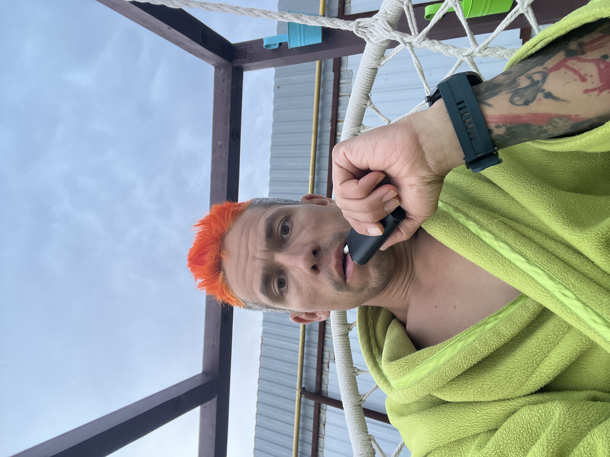 A medium shot depicts a man holding a black electronic device to his mouth while wearing a green bathrobe and a smartwatch. He has short, dyed orange hair on top. The background includes a white macrame-style chair, a metal structure with a partially visible blue container, and a gray corrugated metal wall with yellow pipes. The setting appears to be outdoors with an overcast sky. The location is Chaikovsky, Russia.