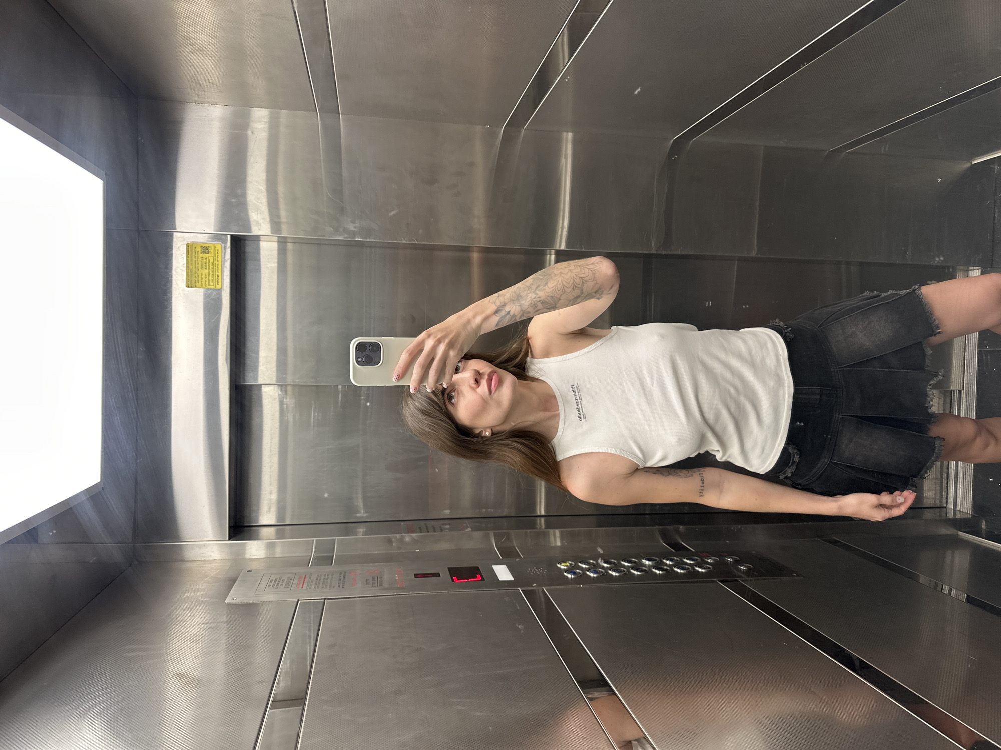 A woman with brown hair is taking a selfie in an elevator with a metal interior. She is wearing a white tank top and a black skirt. The elevator buttons are visible, indicating the selected floor as "1." There is a sticker on the wall next to the door with yellow, white and black text. Given the style of signage, architecture, and clothing, the location could be Tel-Aviv, Israel, but further specifics are unavailable.