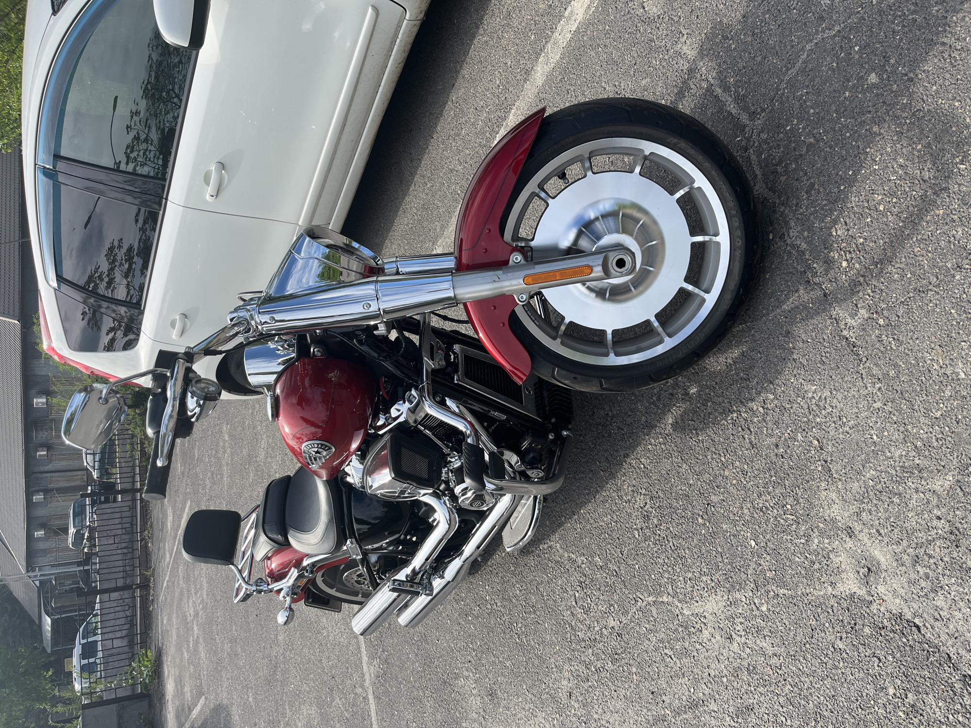 The image depicts a red and chrome motorcycle parked on an asphalt surface, possibly a parking lot or street. A white car is partially visible to the left of the motorcycle's front. The environment includes a building and greenery in the background. It is not possible to determine if this is in Kryvyi Rih, Ukraine with the information provided.