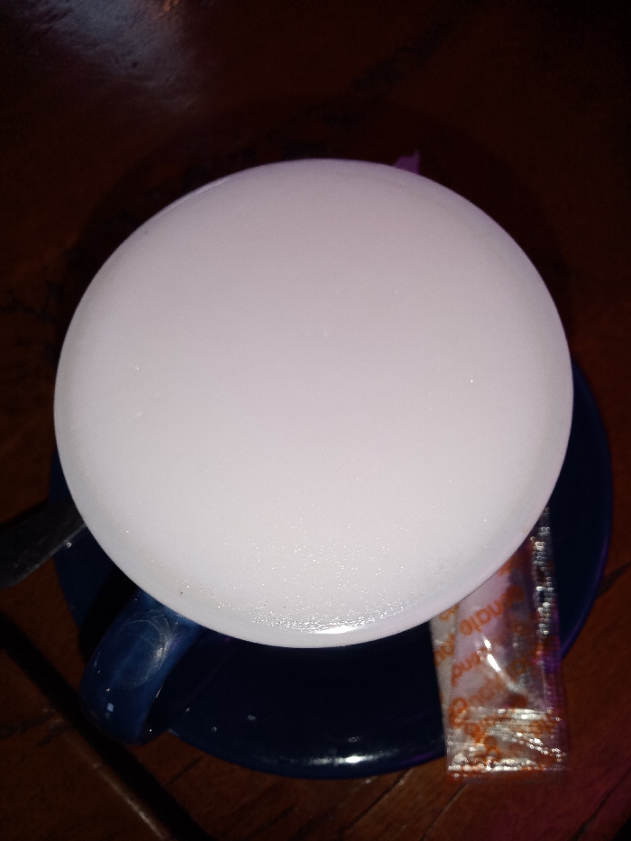 A top-down, slightly angled shot captures a hot beverage served in a dark blue cup on a matching saucer, resting on a dark wooden table. The drink features a thick, bright white, frothy layer, characteristic of a cappuccino or latte, filling the entire visible surface of the cup. To the left, a silver spoon or stirrer is partially visible, resting on the saucer. To the right, a small, transparent rectangular packet, likely containing a condiment like sugar or creamer, lies on the saucer. Part of the text "mate" and "pind" (possibly from "Indomate" or similar branding) is visible on the packet, which has an orange-brown pattern. The lighting is dim, casting a warm glow on the frothy surface while the surroundings remain dark, suggesting an indoor setting such as a cafe or restaurant, likely during the evening or night. No people are visible, and the scene is serene, focused solely on the prepared drink.