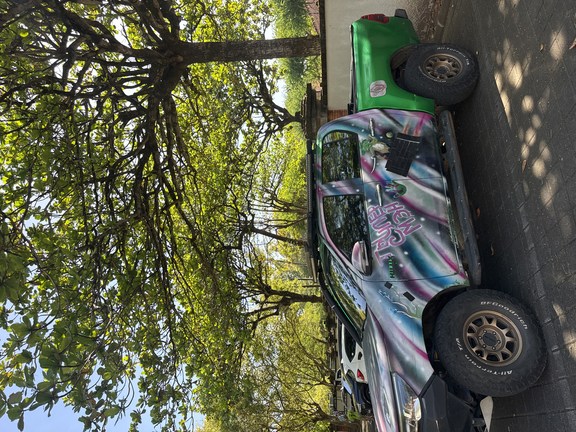 The image shows a parking area in Kuta Selatan, Indonesia, lined with trees that cast shadows on the brick-paved ground. A parked, customized green and painted pick-up truck with large tires takes up the primary view. The truck's paint features the word "HILUX" and other graphics. Other parked vehicles are partially visible behind the truck. A wall and some foliage separate the parking area from what seems like a residential zone in the background. The sky is partly visible through the dense tree canopy.
