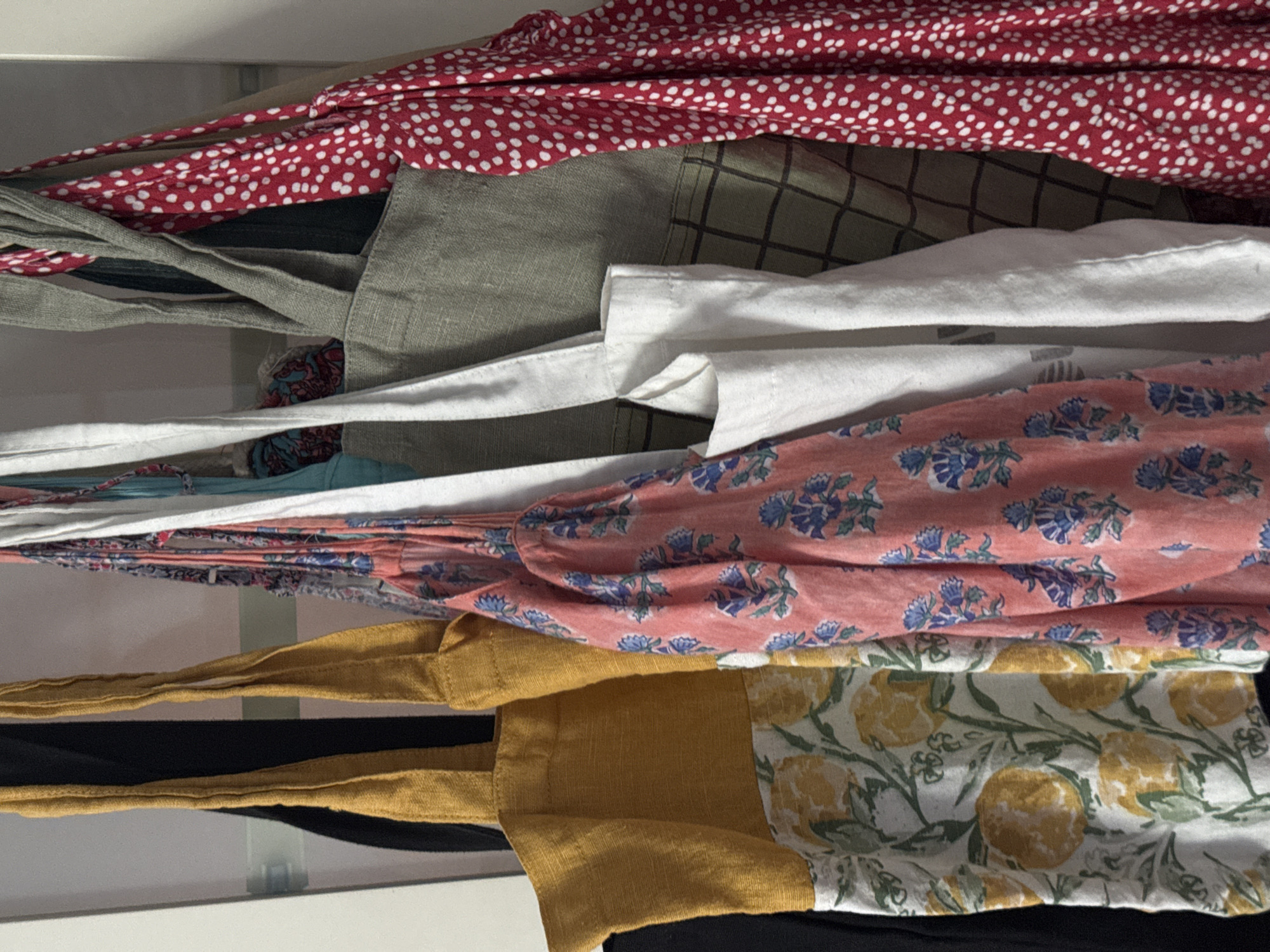 The image shows a close-up of a drawer filled with various reusable shopping bags. At the top is a red bag with white polka dots. Below that is a grey-green bag with a plaid pattern. Underneath that is a white bag. Next is a coral-colored bag with a floral pattern. At the bottom is a yellow bag with a floral pattern and a black bag. The bags are stacked and appear to be compressed within the drawer. The scene is indoors. There is no evidence of the location being Kazan, Russia, or any information about people, subjects, actions, or interactions in the provided image.